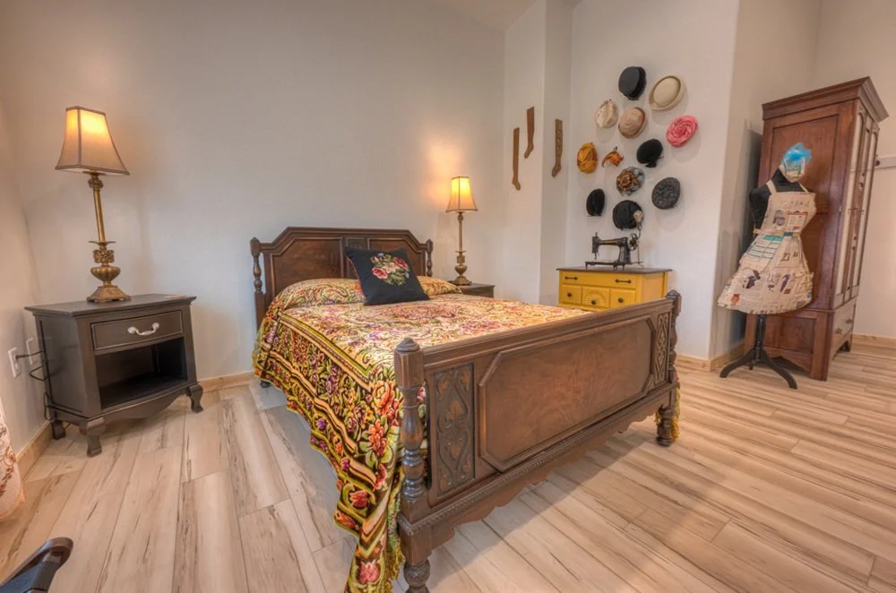 A bedroom with a wooden bed, floral bedding, and a decorative pillow. Two nightstands with lamps on each side. Wall decor includes hats and socks. A yellow cabinet with a vintage sewing machine, a wooden wardrobe, and a mannequin dressed in fabric.
