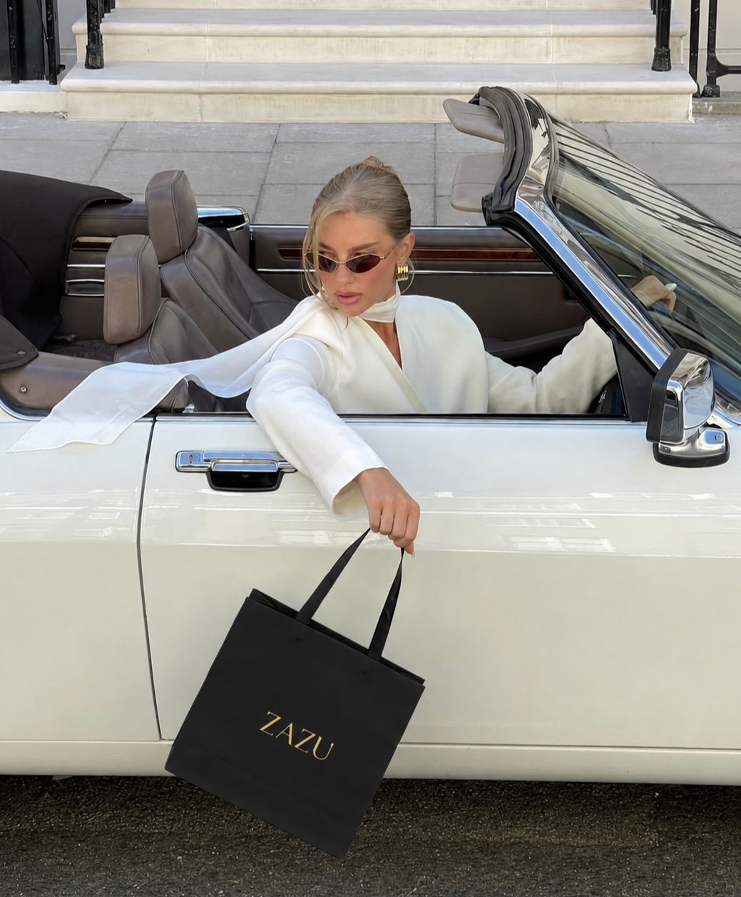 A woman with blonde hair, sunglasses, and gold earrings sitting in a white convertible car, holding a black shopping bag with the brand name ZAZU, parked in front of a building with steps.