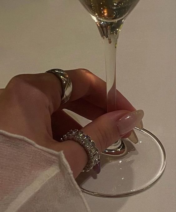 Close-up of a person's hand holding a glass of white wine, with rings and painted nails, against a plain background.