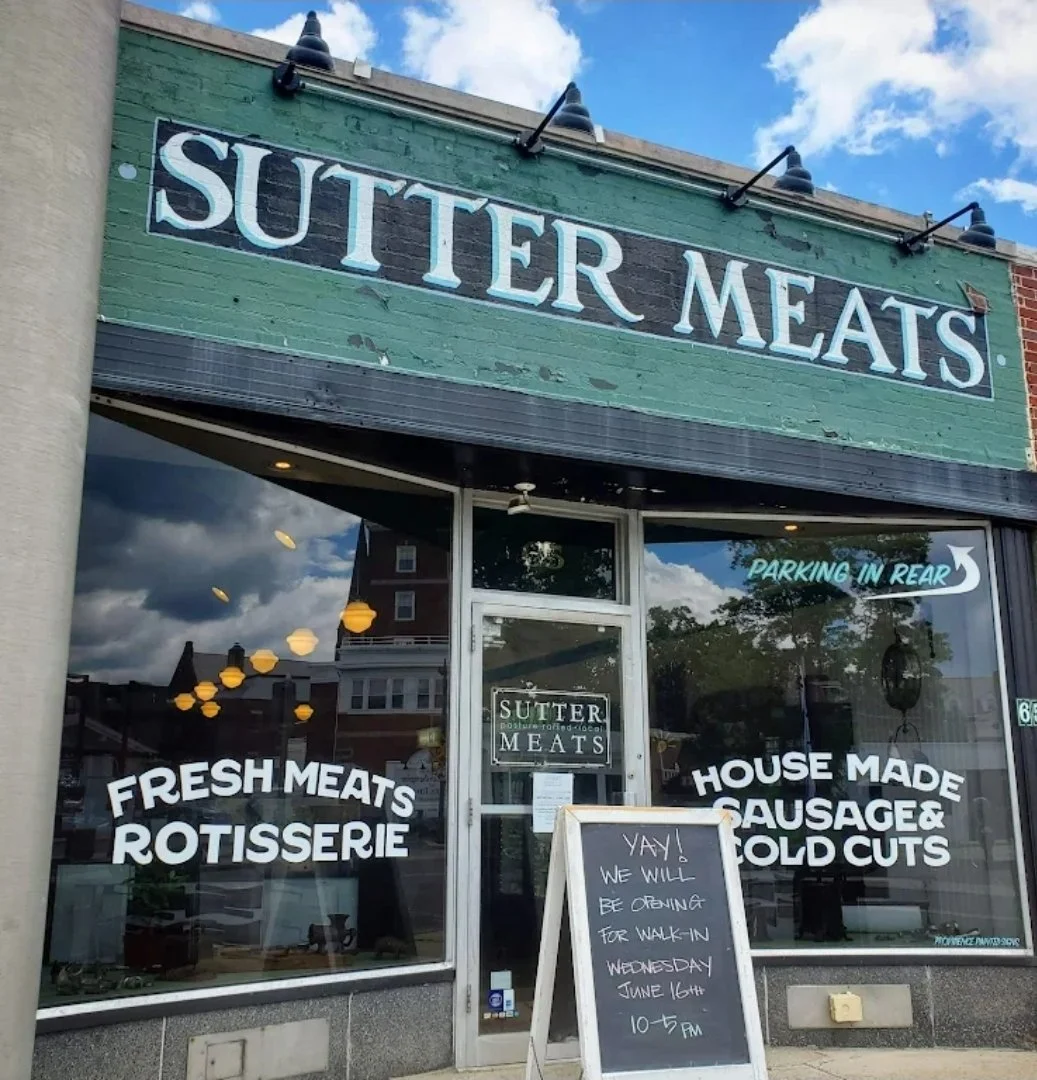 The exterior of Sutter Meats, a butcher shop with a green painted brick facade. The large sign reads 'SUTTER MEATS'. The shop window has white lettering advertising 'FRESH MEATS ROTISSERIE' and 'HOUSE MADE SAUSAGE & COLD CUTS'. A chalkboard sign in front announces the opening for walk-in customers on Wednesday, June 16th, from 10 am to 7 pm. There is also a decal on the window indicating parking in the rear. Reflections of trees and buildings can be seen in the glass.