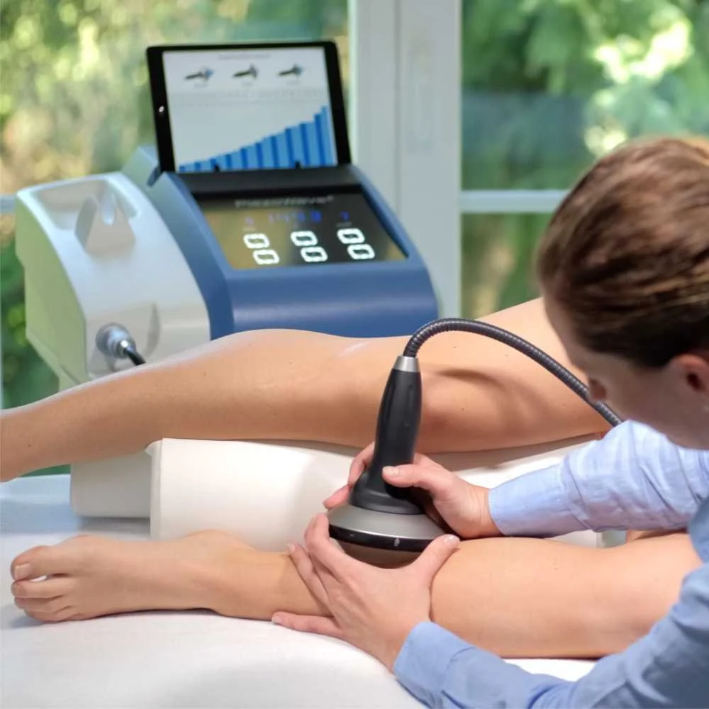 Medical technician uses ultrasound device on patient's leg in a clinical setting with a digital monitor showing graphs and charts.