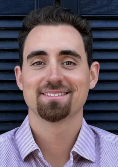 A smiling young man with dark hair and a goatee, wearing a light purple collared shirt, standing in front of a dark blue louvered wall.