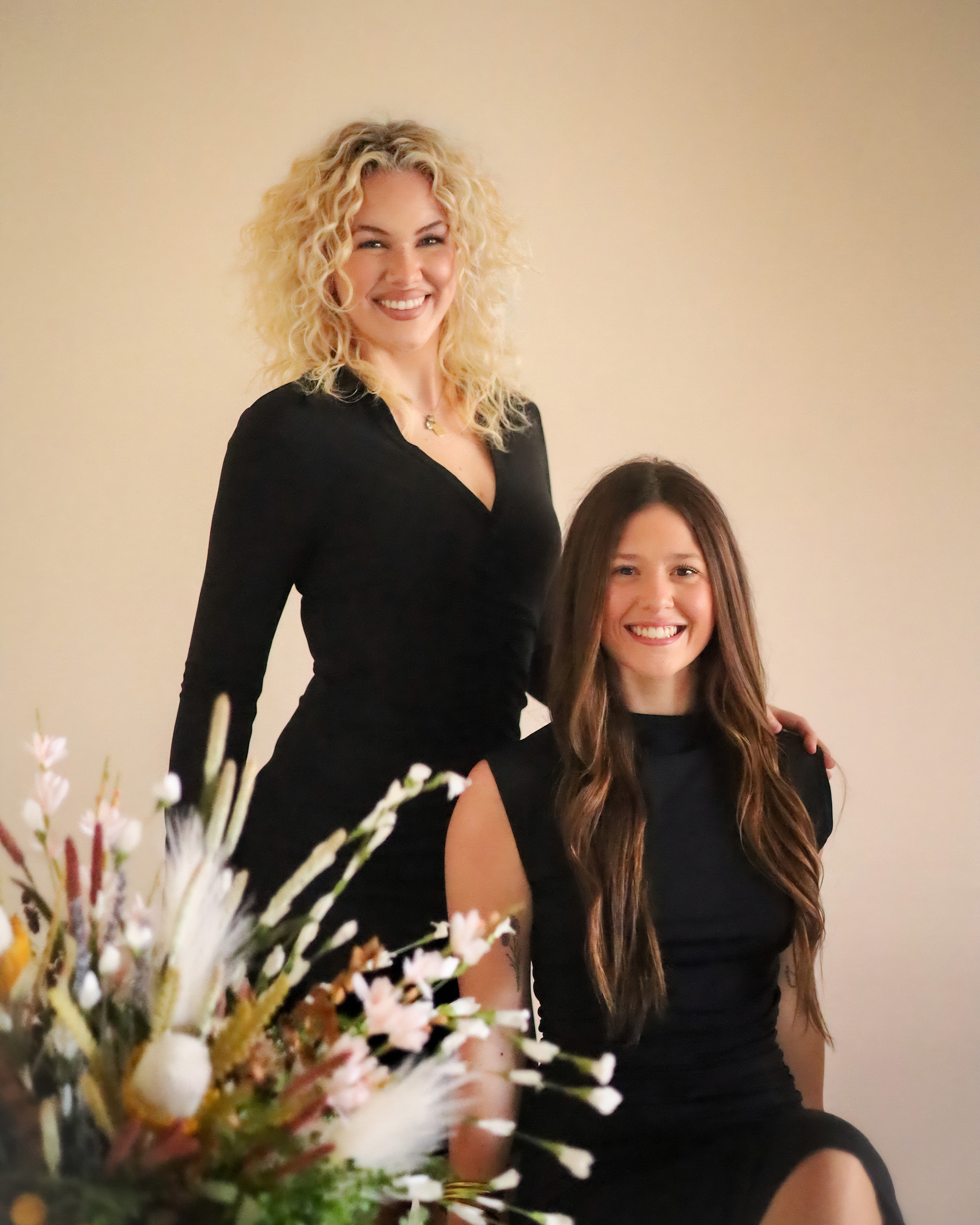 A smiling woman with curly blonde hair standing next to a young girl with long straight brown hair. They are both dressed in black and are indoors with a plain light-colored wall background. A flower arrangement with white blossoms is in the foreground.