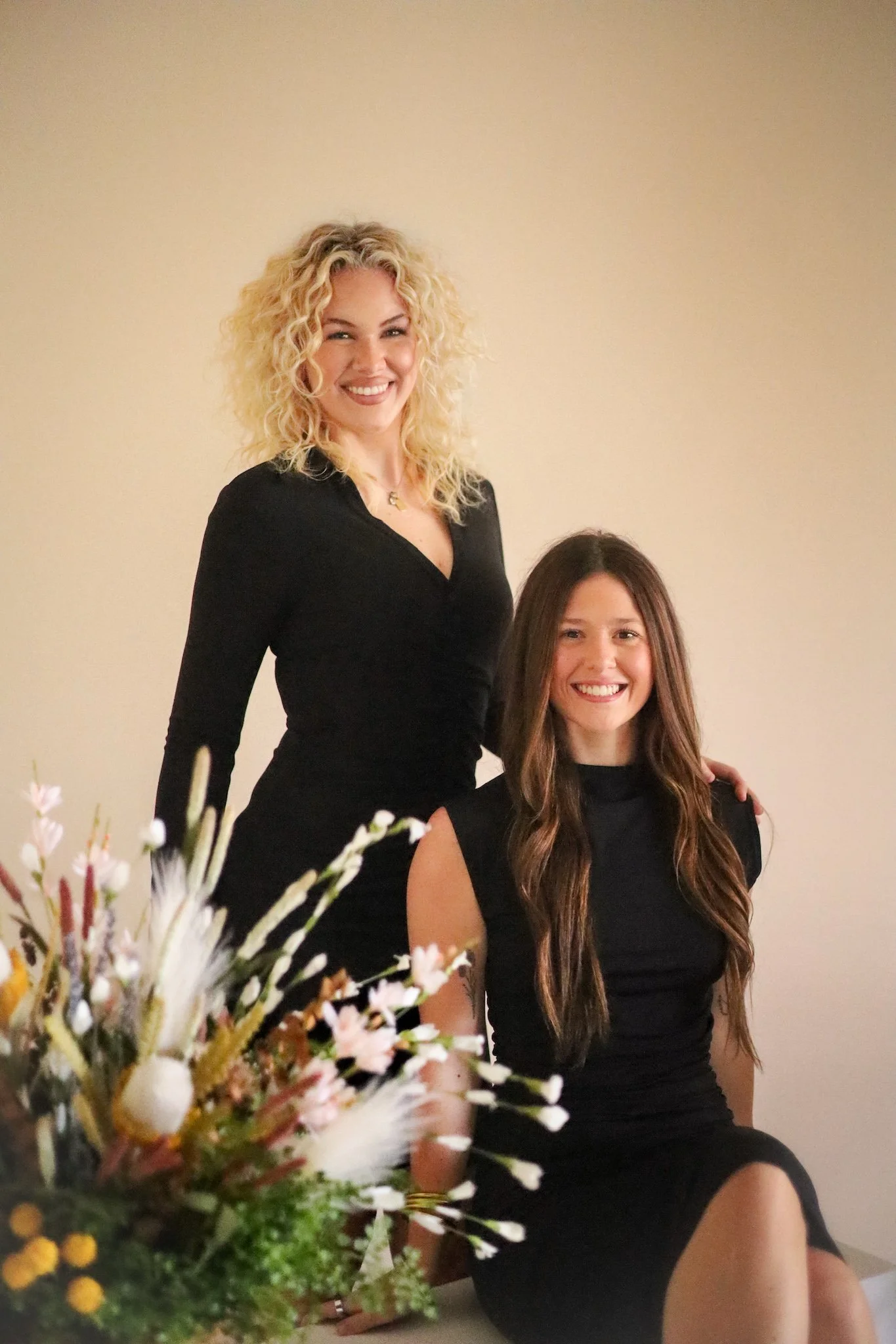 A woman with curly blonde hair and a woman with long straight brown hair, both smiling, dressed in black, sitting indoors in front of a plain beige wall. A bouquet of flowers is visible in the foreground.
