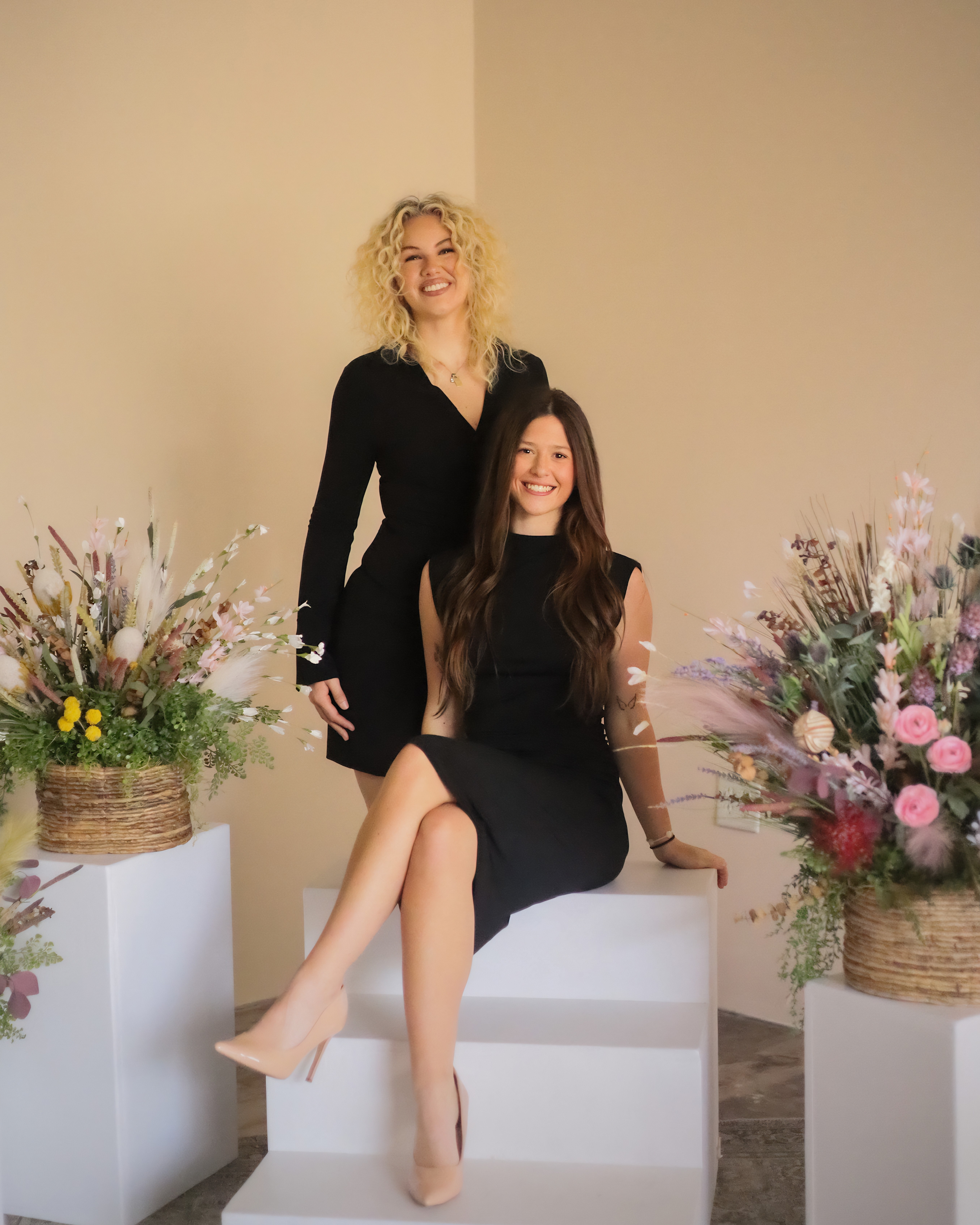 Two women, one with curly blonde hair and the other with long brown hair, posing among floral arrangements, smiling at the camera.