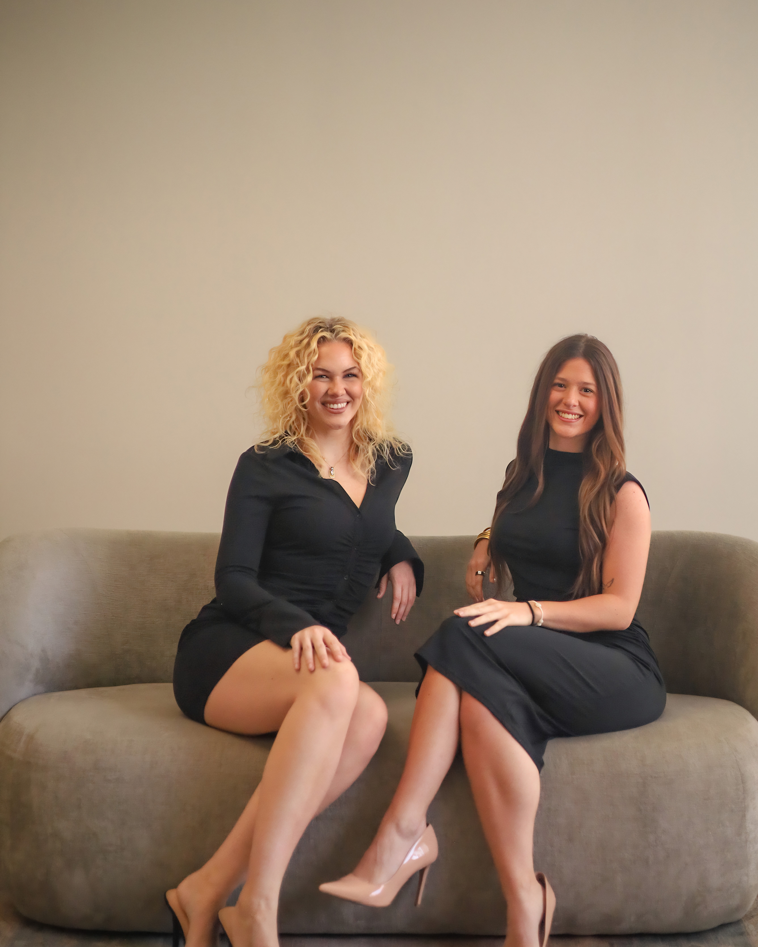 Two women sitting on a gray couch smiling at the camera, wearing black dresses, with one woman wearing nude high heels and the other wearing black heels.