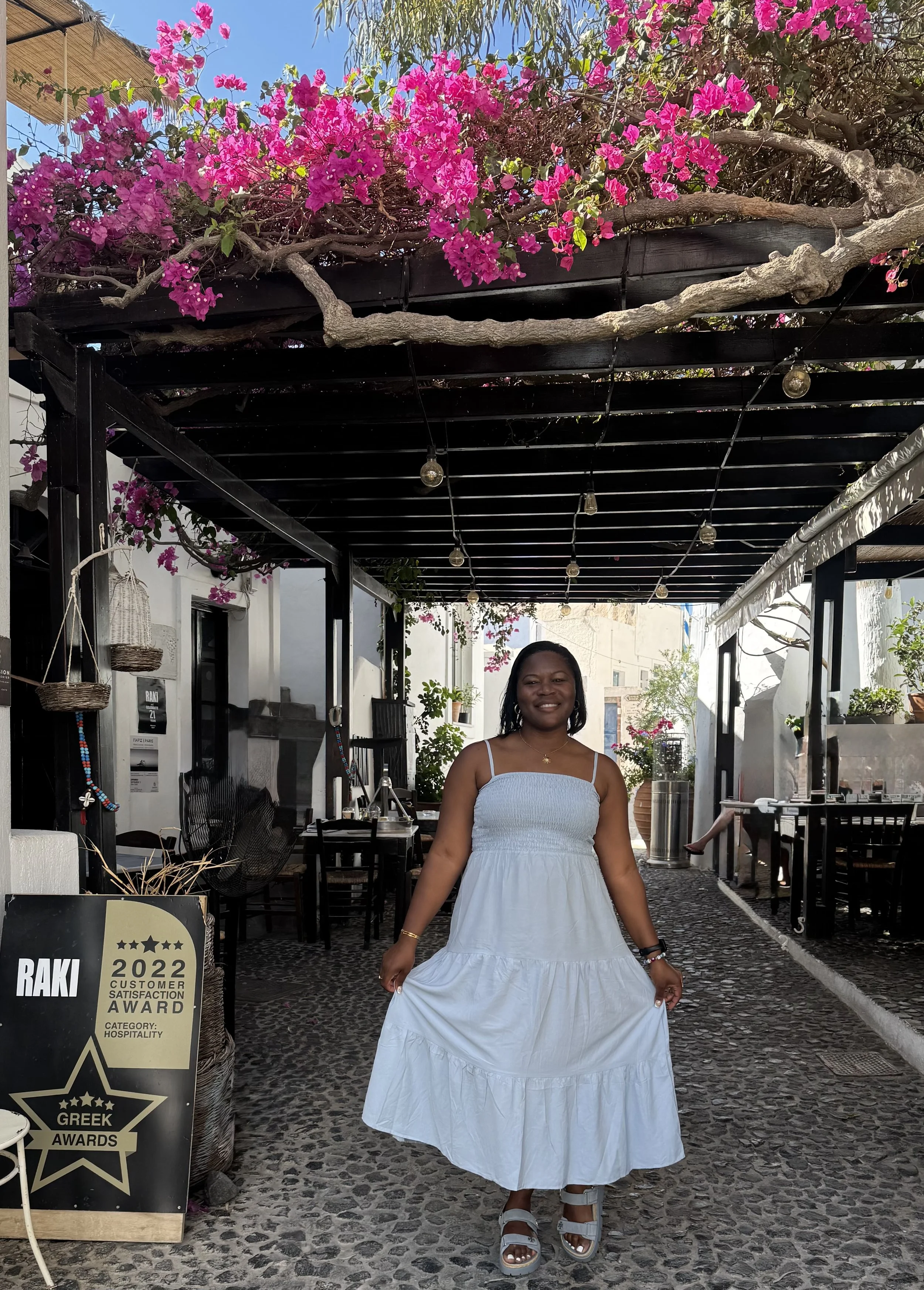 Woman in a white dress standing outdoors under a wooden pergola with pink bougainvilla flowers and string lights, smiling at the camera.