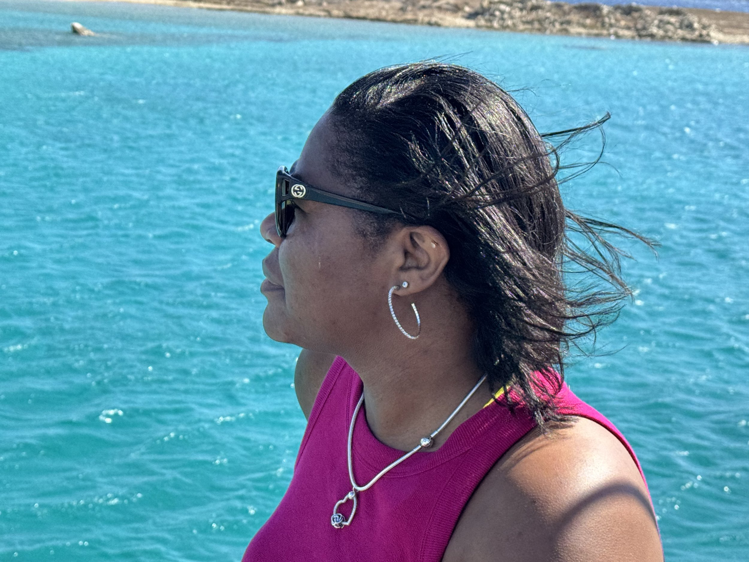 Leona Partis side profile picture in Greece wearing sunglasses and ear jewelry, near a body of blue water with rocky shoreline in the background.