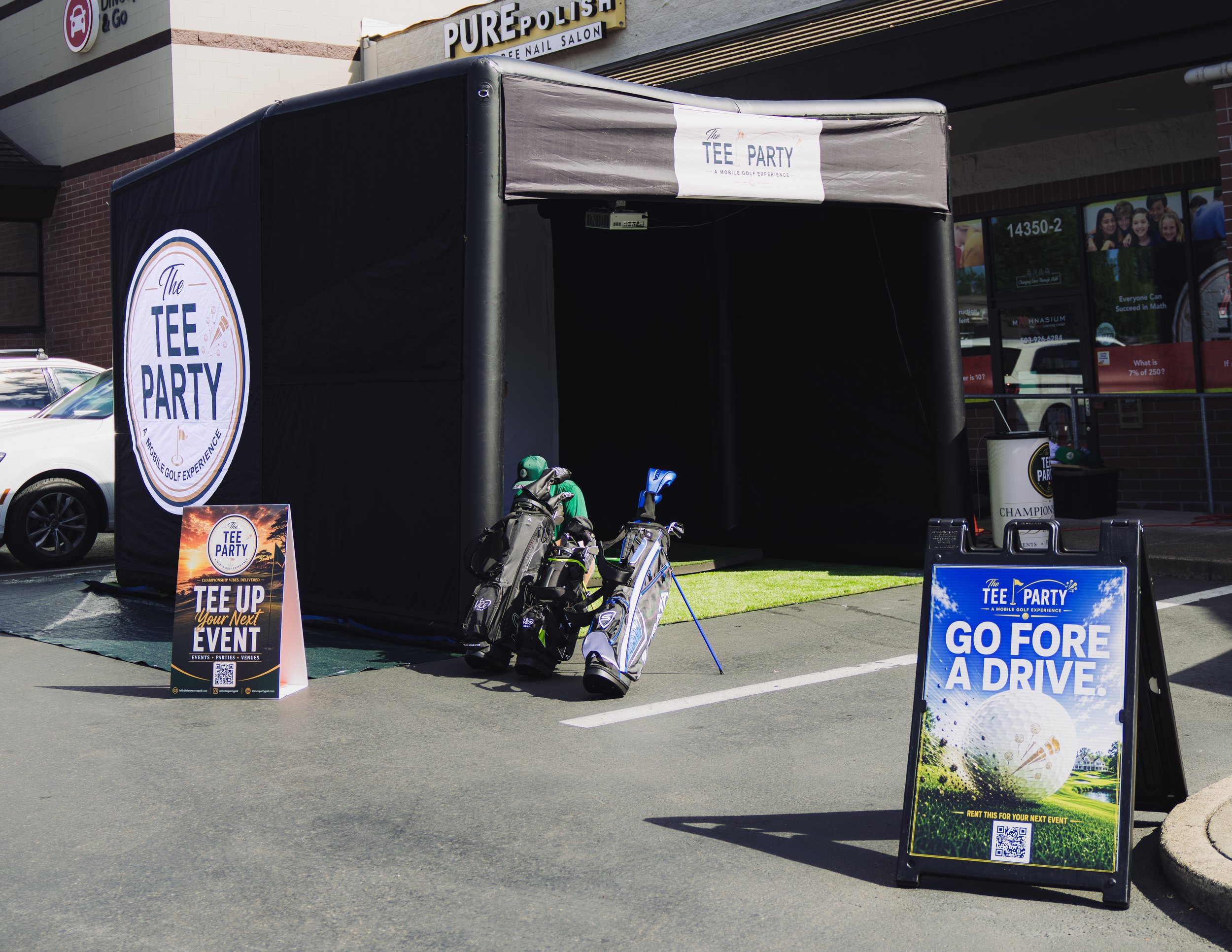 A black canopy tent advertising a golf event called 'The Tee Party' with various signage promoting the event, including a sign that says 'Go for a drive' and one that says 'Rent this for your next event' with a QR code, set up outside a building with a sign that reads 'Pure Polish' and a window showcasing a group of children.