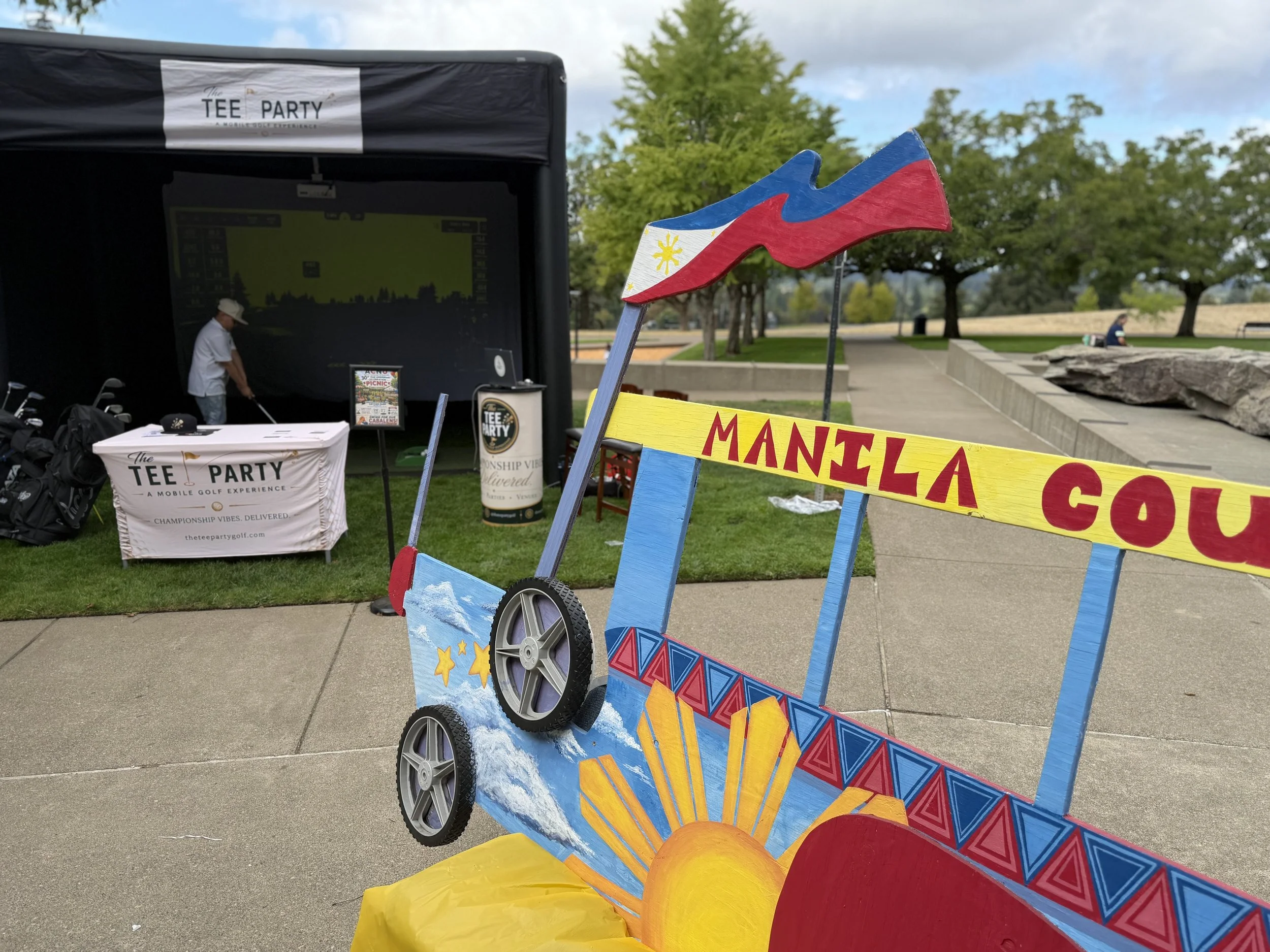 Colorful decorated cart in front of a green park with trees. In the background, a black tent with a sign for 'The Tee Party' golf event and a person preparing golf clubs.