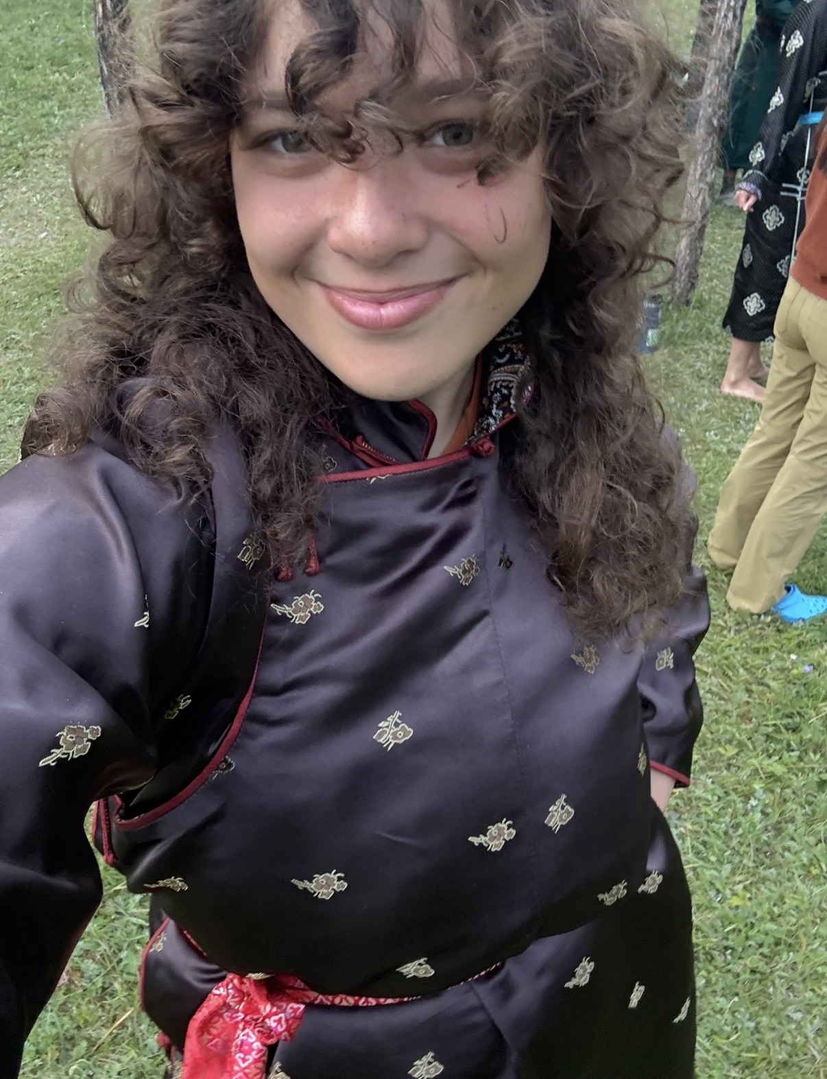 A person with long, curly hair is taking a selfie outdoors. They are wearing a black deel with gold embroidery and a red patterned sash at the waist. In the background.