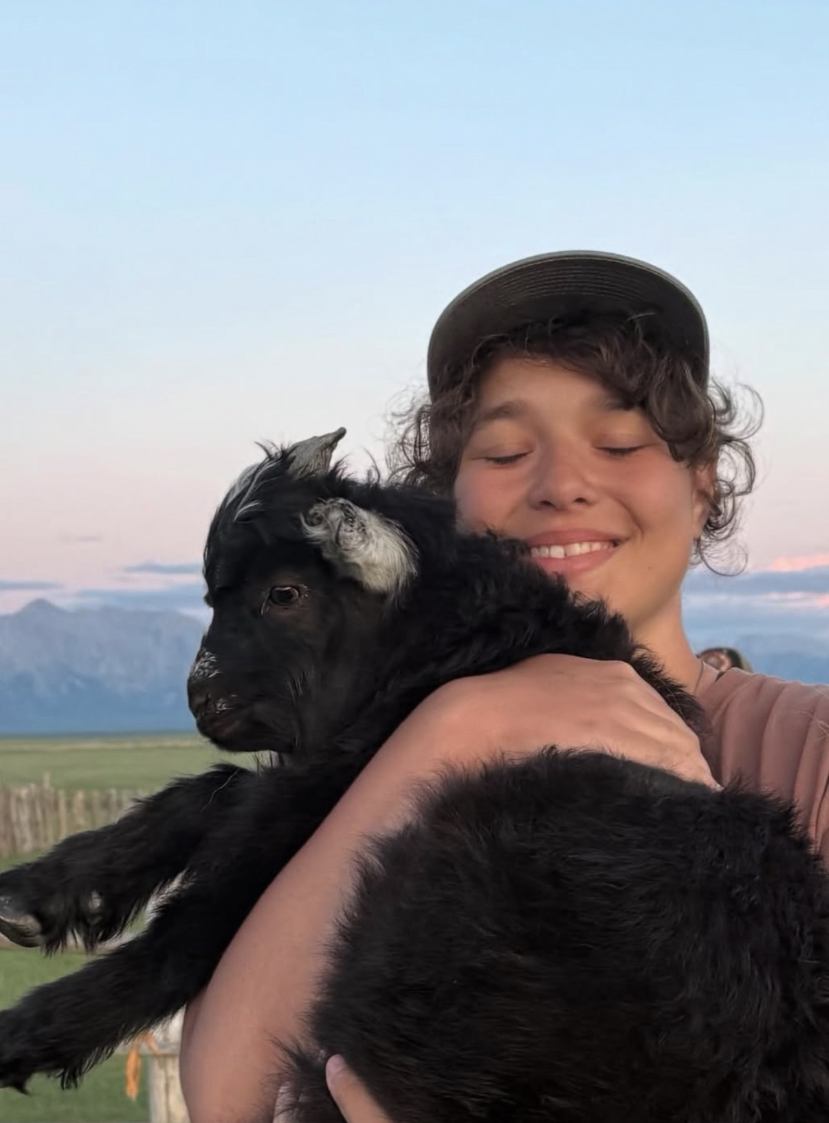 A person with curly hair wearing a baseball cap hugging a black and white goat outdoors with mountains in the background