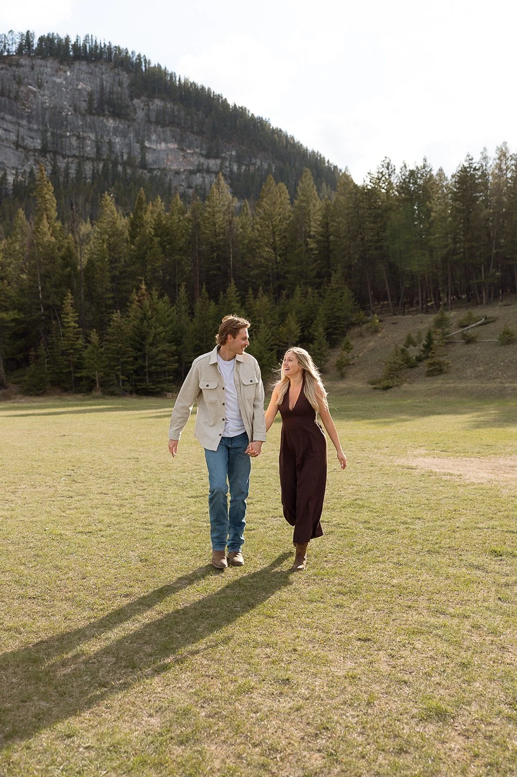 A young couple walks hand in hand across a grassy field, surrounded by a forest and mountain landscape, enjoying a sunny day.