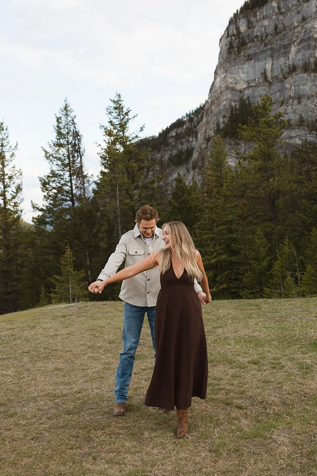 A couple dancing and enjoying in a grassy field with pine trees and a mountain in the background.