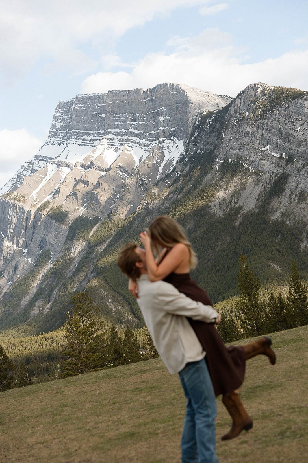 A man lifting a woman in the air during daytime with a large mountain range in the background and trees in the foreground.
