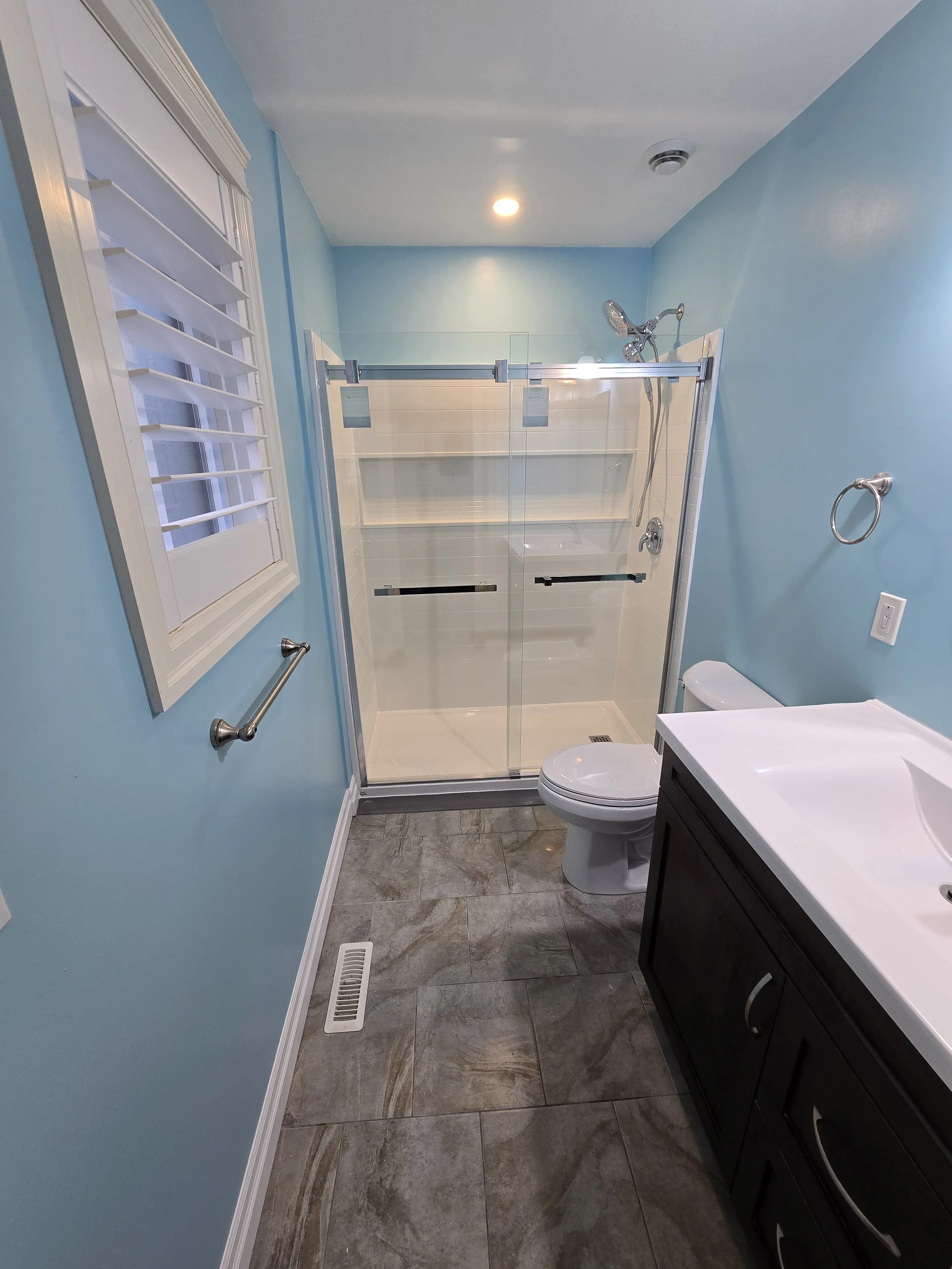 Acrylic shower inster:
Bathroom with light blue walls, a window with white shutters, a glass-enclosed shower, a white toilet, a dark vanity with a white sink, a towel ring, a wall vent, and ceramic tile flooring.