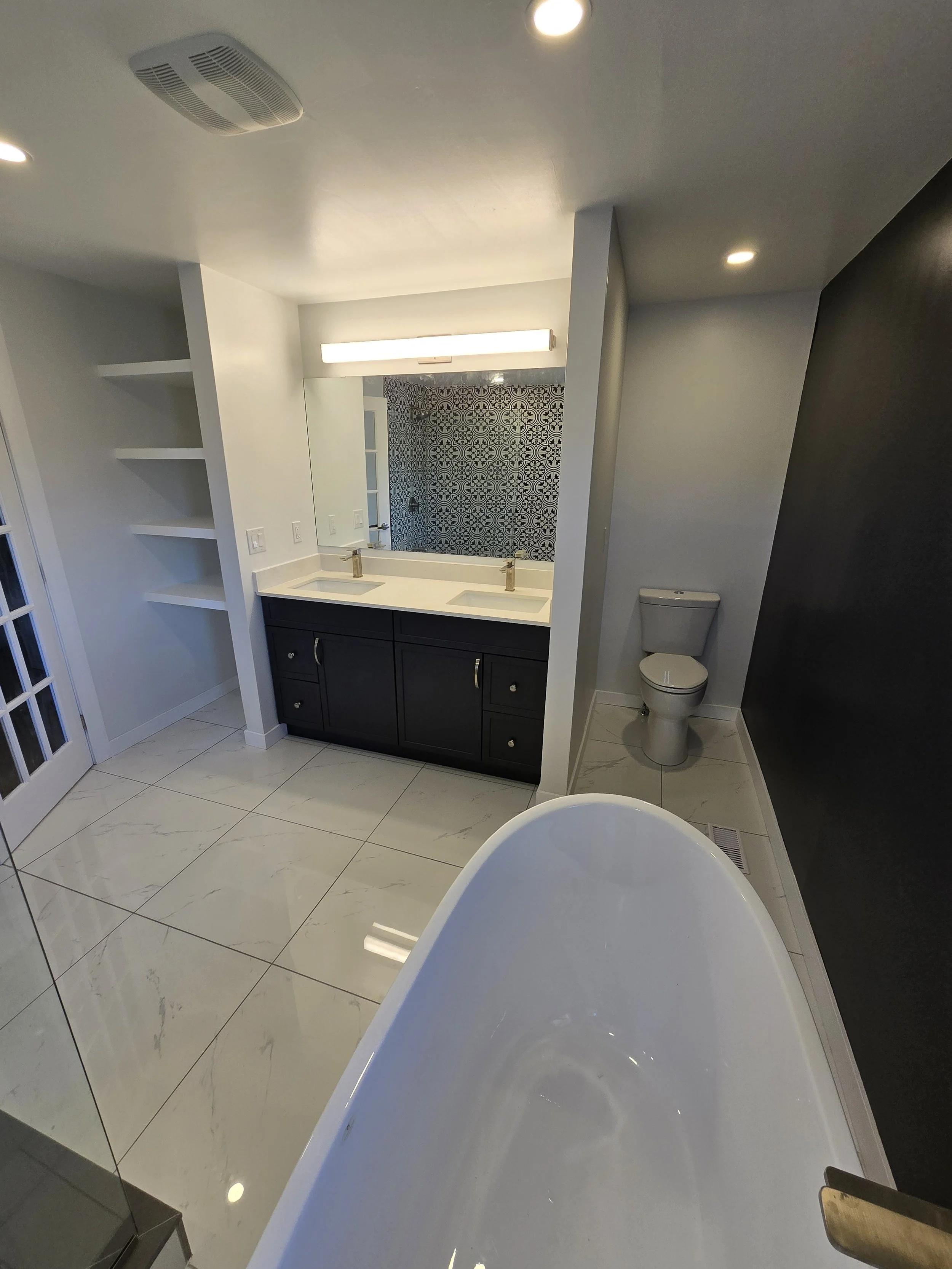 A bathroom with white tiled flooring, a free-standing bathtub, a vanity with a large mirror, a patterned tile wall in the shower, and a visible toilet. Recessed ceiling lights illuminate the space.