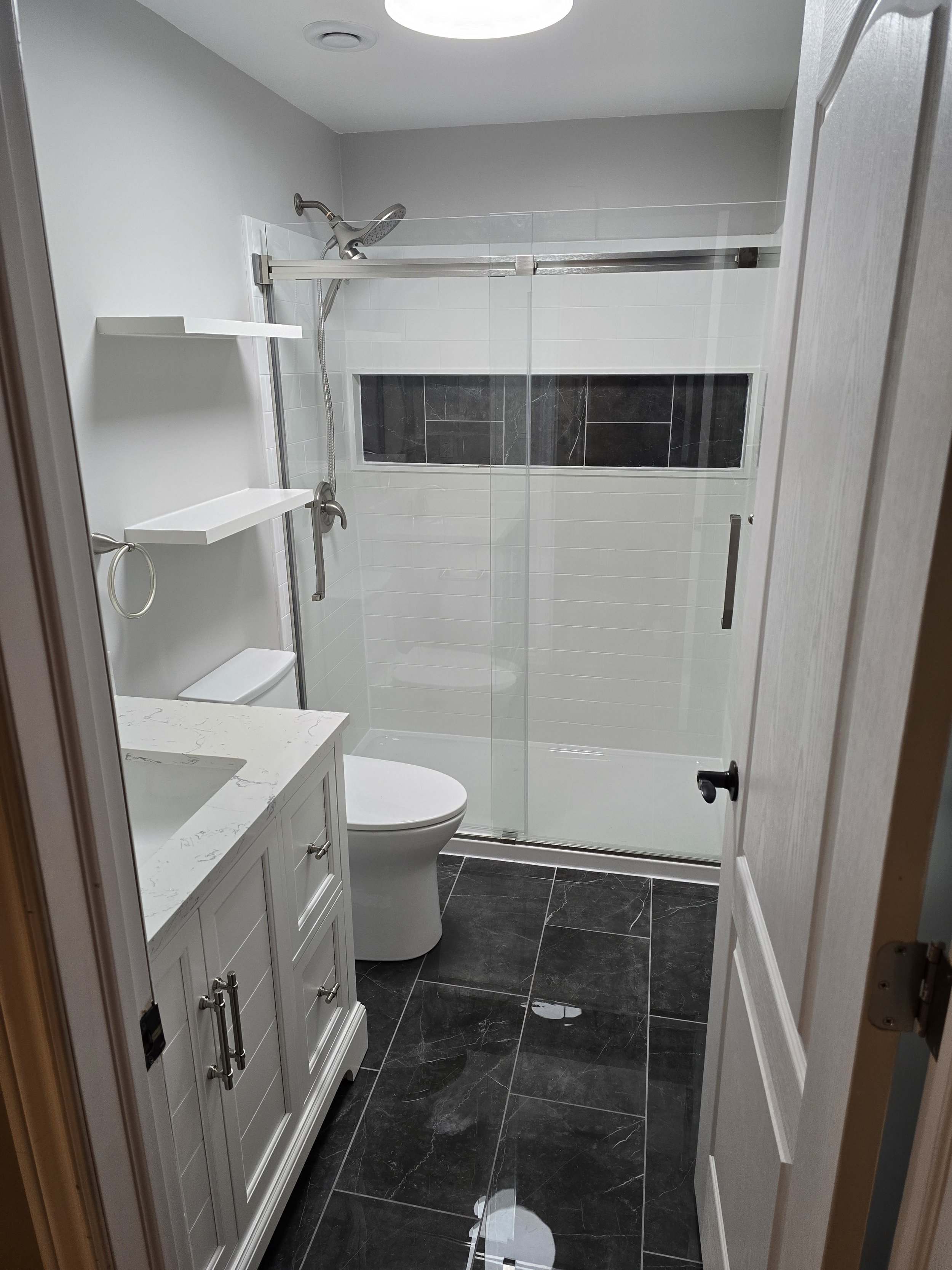 After
Modern bathroom with black marble floor tiles, a white vanity with multiple drawers, a toilet, a glass-enclosed shower with white and black tiles, and a small built-in shelf in the shower wall.