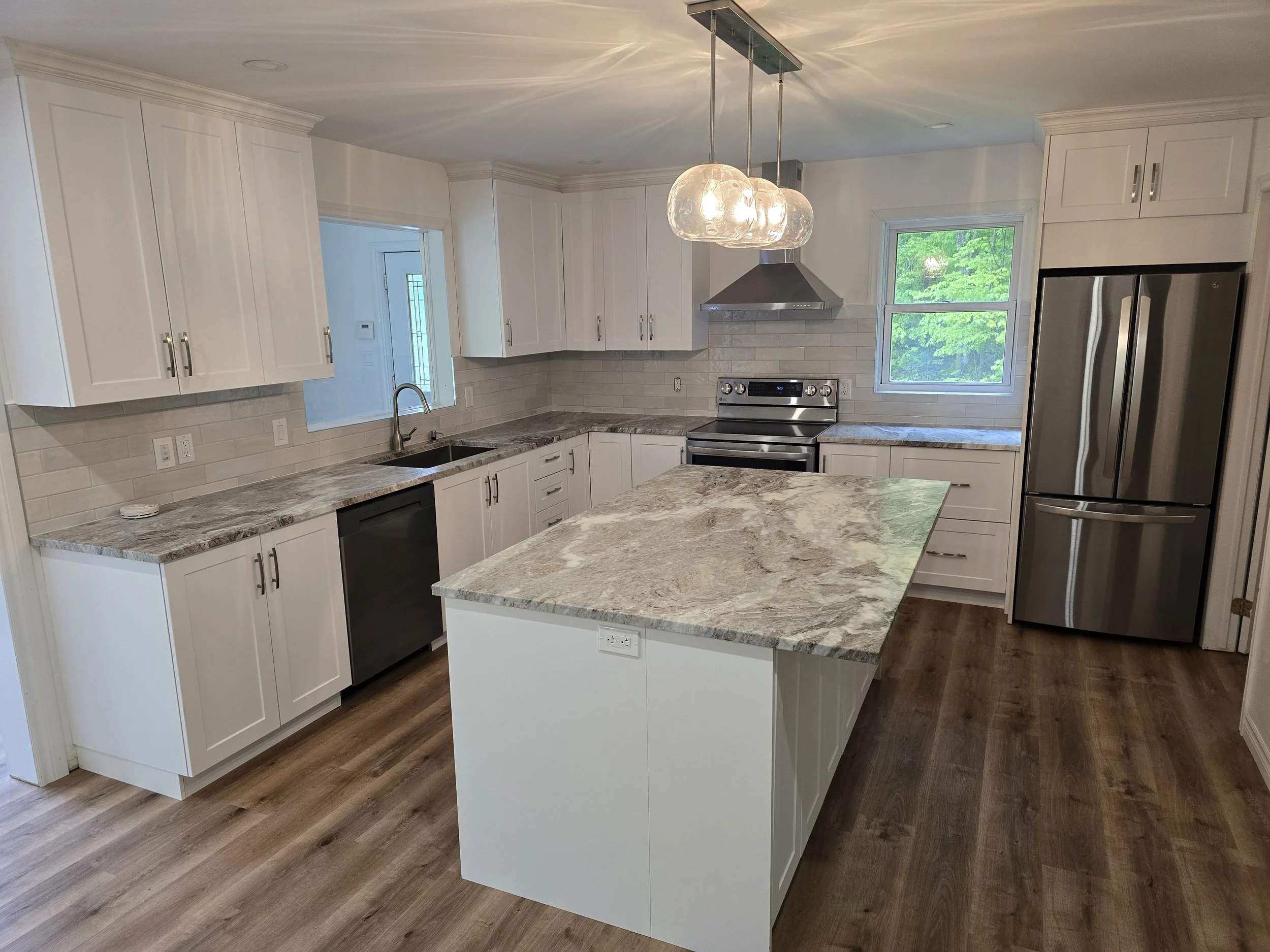 A modern kitchen with white cabinets, a marble countertop island, stainless steel appliances including a refrigerator and oven, a range hood, a sink beneath a window, and a hanging light fixture.