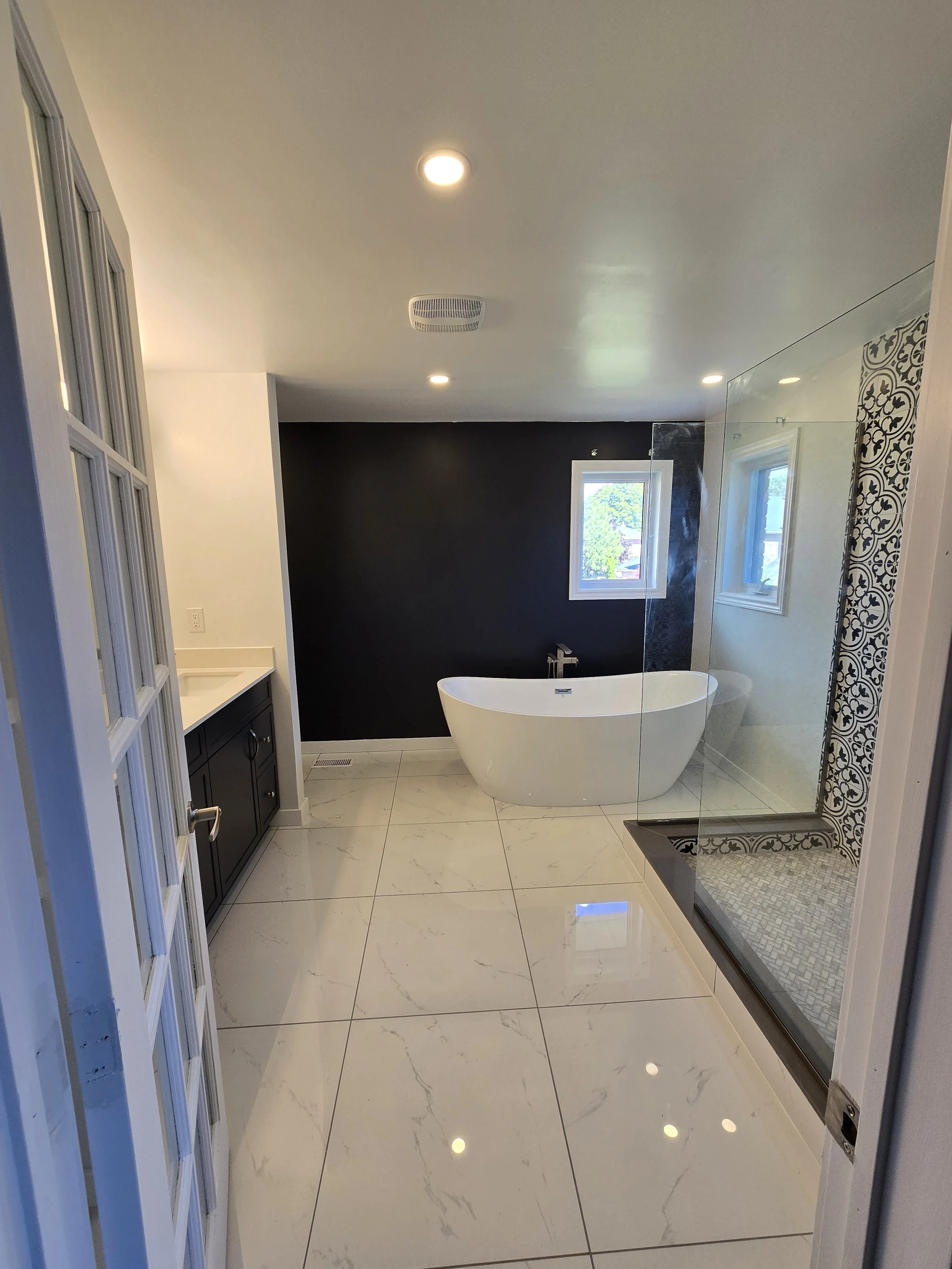 Modern bathroom with white marble floor and a freestanding bathtub in front of a dark accent wall, with large windows and a glass-enclosed shower with patterned tiles.