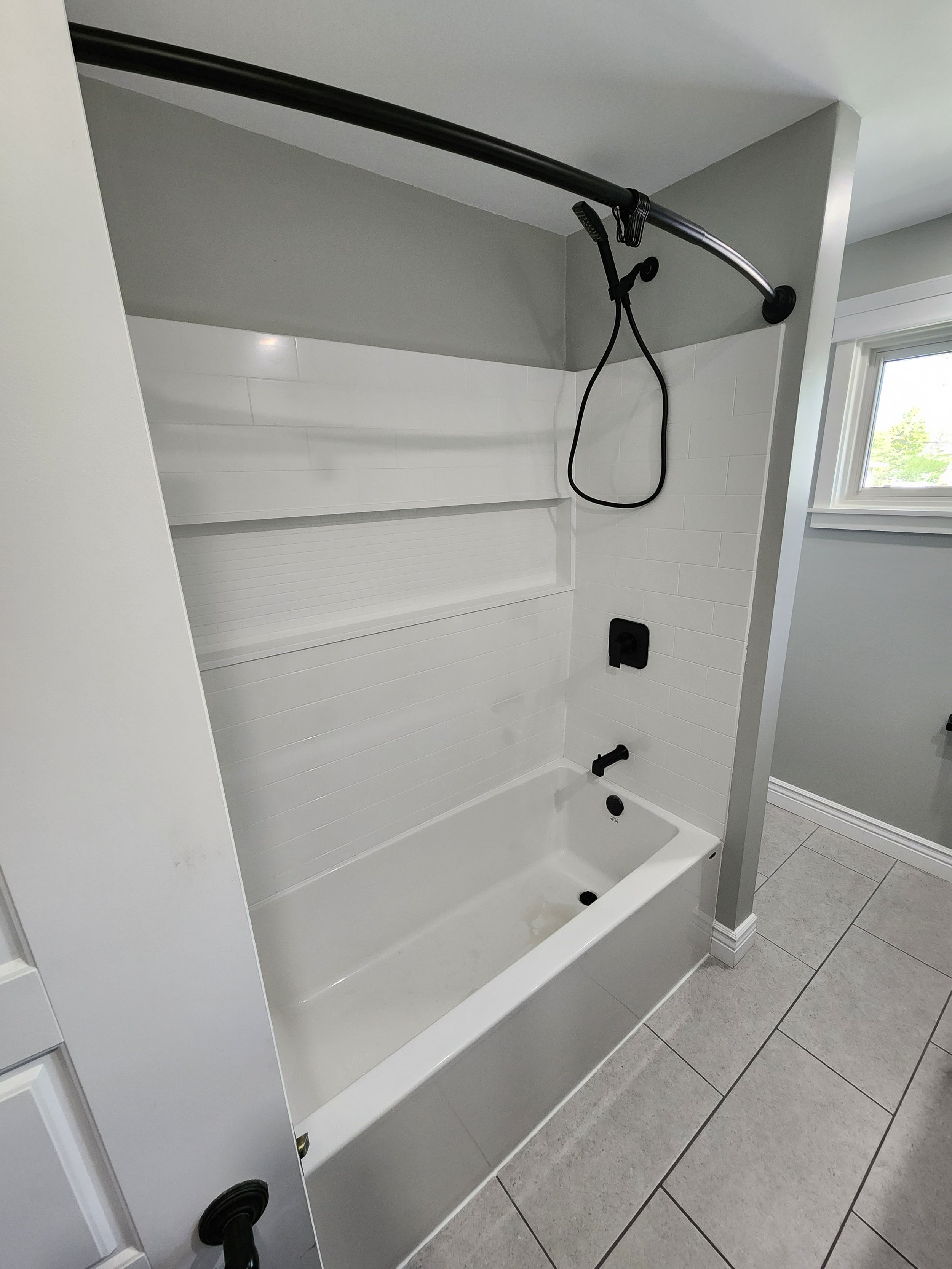 Acrylic shower insert:
A white bathtub with a black faucet and a black showerhead. The walls are white with a built-in shelf. The bathroom has gray tiles and a window with a view of trees outside.