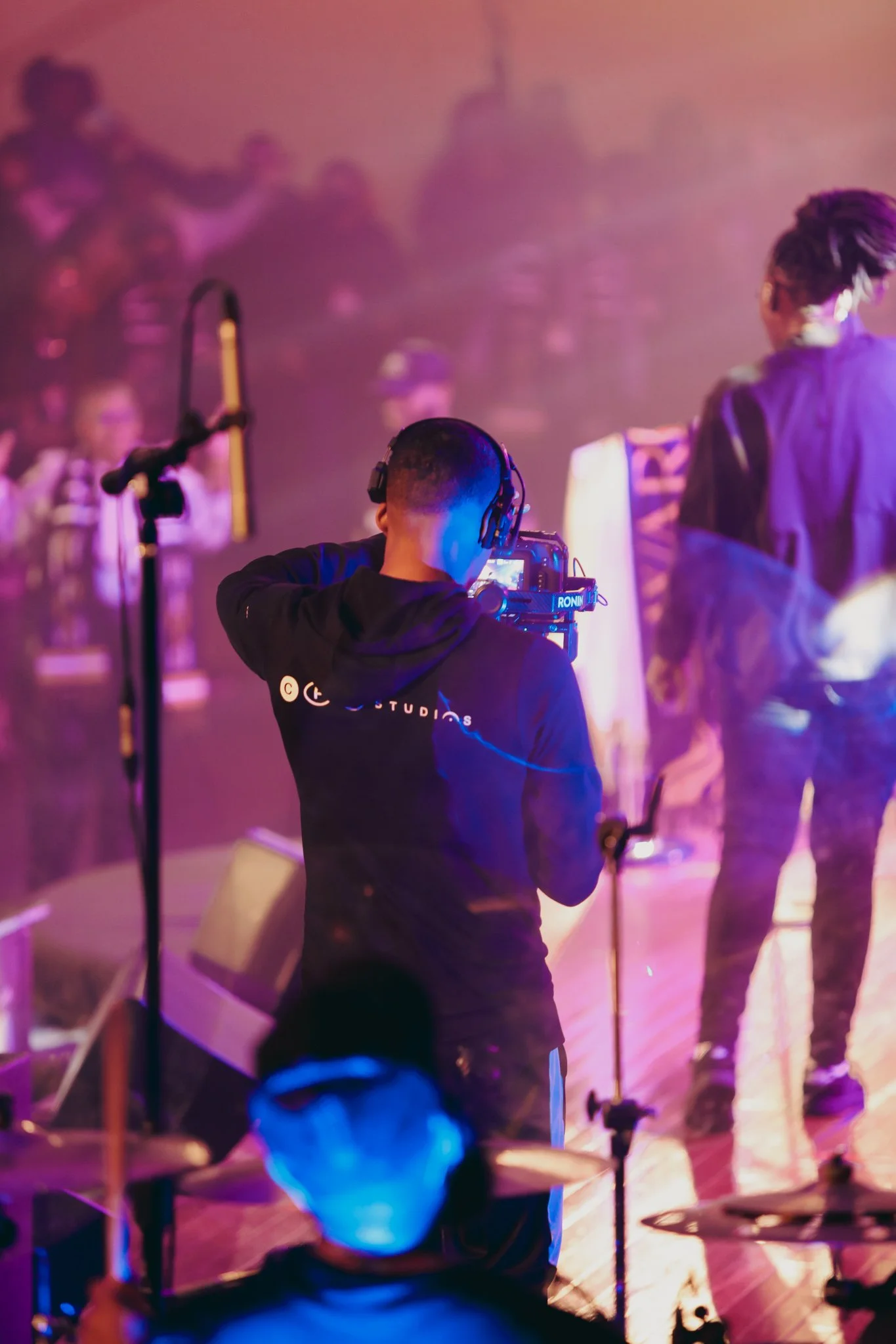 A person filming a band with a professional camera at a live music event, with musicians playing on stage in the background, illuminated by colorful stage lights.
