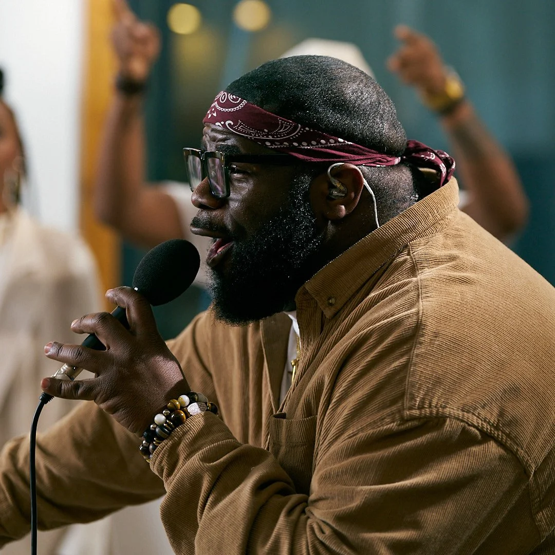 A man wearing glasses and a red bandana singing into a microphone.