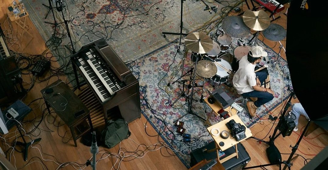 A musician sitting on the floor with a drum set and musical equipment in a studio or rehearsal space.