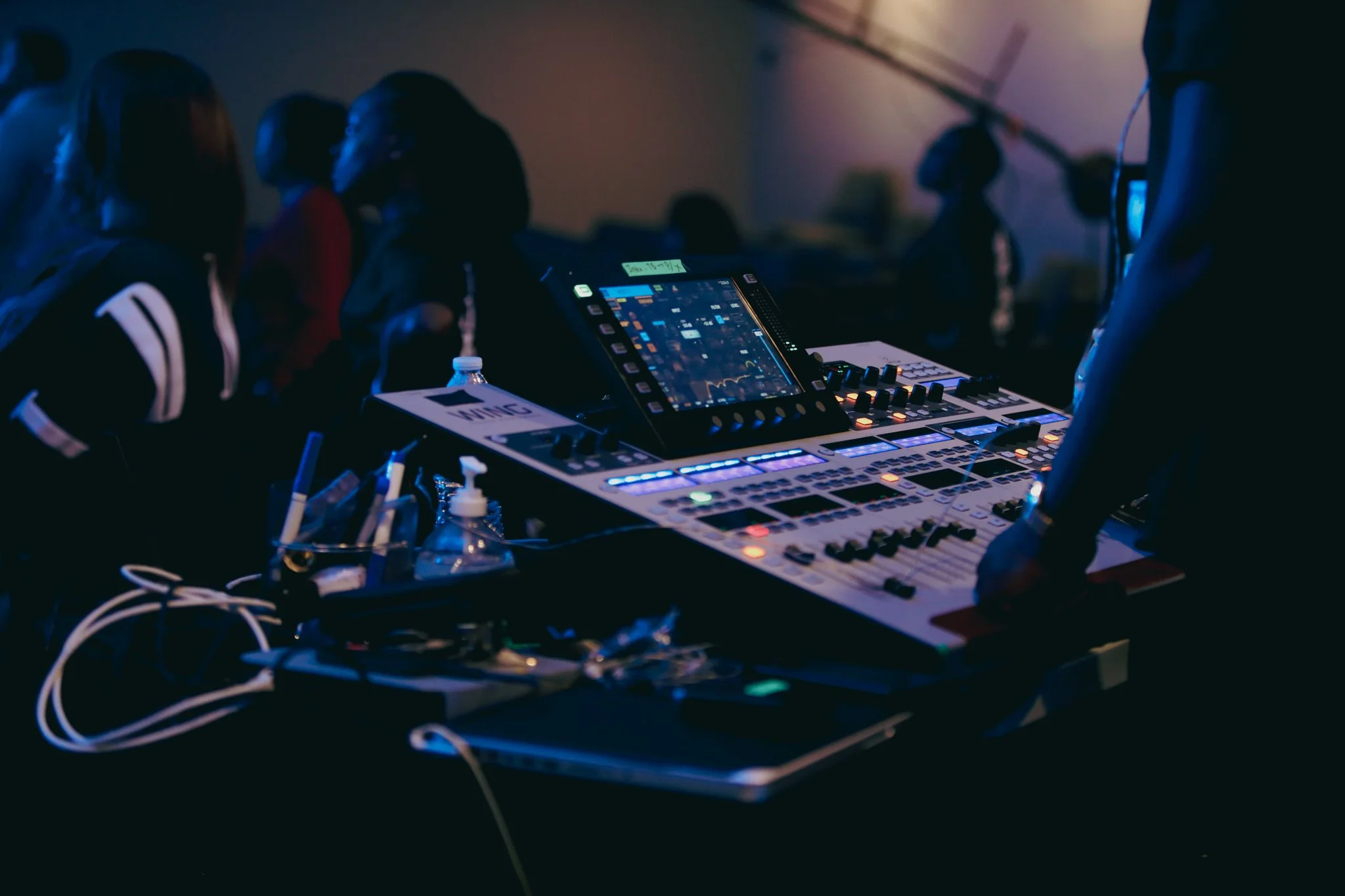 A DJ equipment setup with electronic music controllers and a touchscreen display, surrounded by people in a dark environment, possibly at a concert or club.