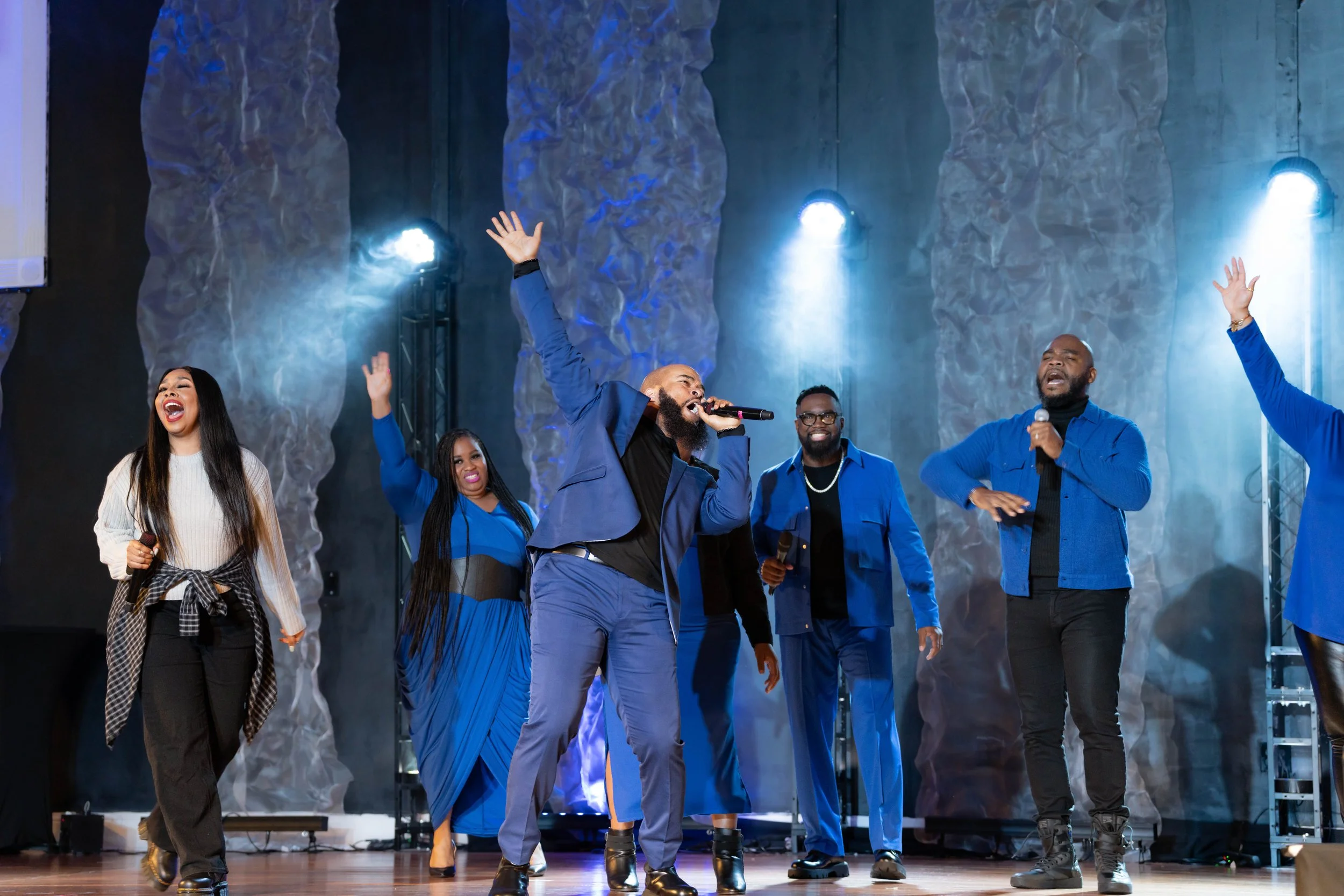 A diverse group of people on stage, some dancing and singing with microphones, with expressions of joy, in front of textured blue walls and bright stage lights.