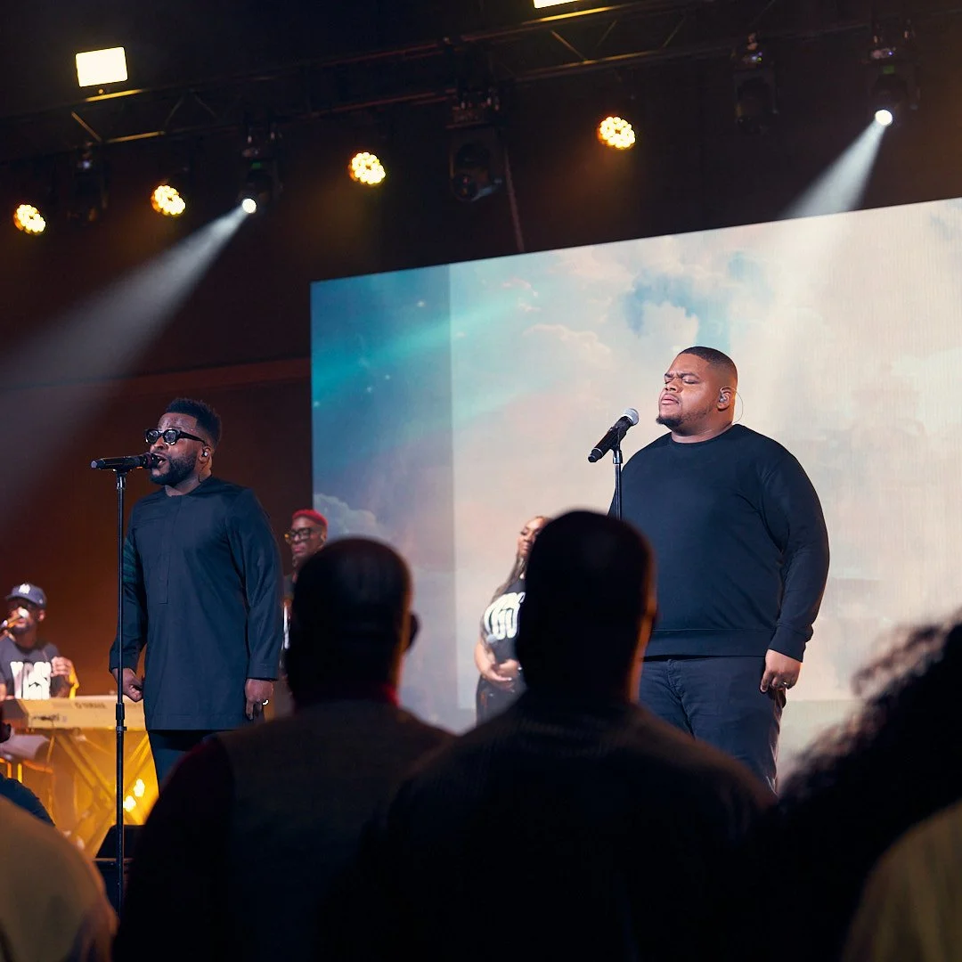 Two men singing on stage with microphones, one wearing sunglasses and a black outfit, the other with eyes closed and wearing a black sweatshirt, with a band and audience present.