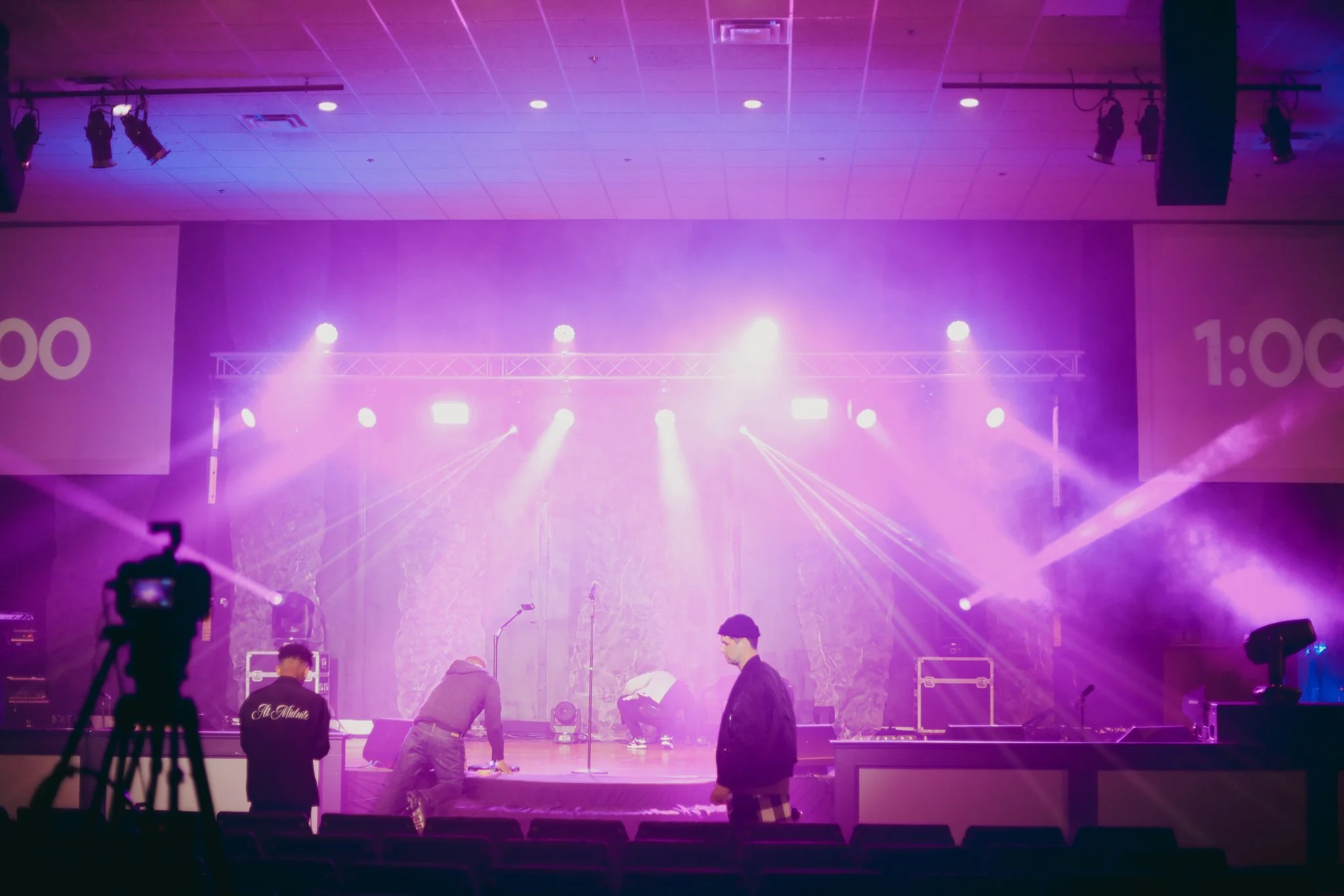 Stage with purple and pink lighting, musical instruments, and crew preparing for a performance in an indoor venue.