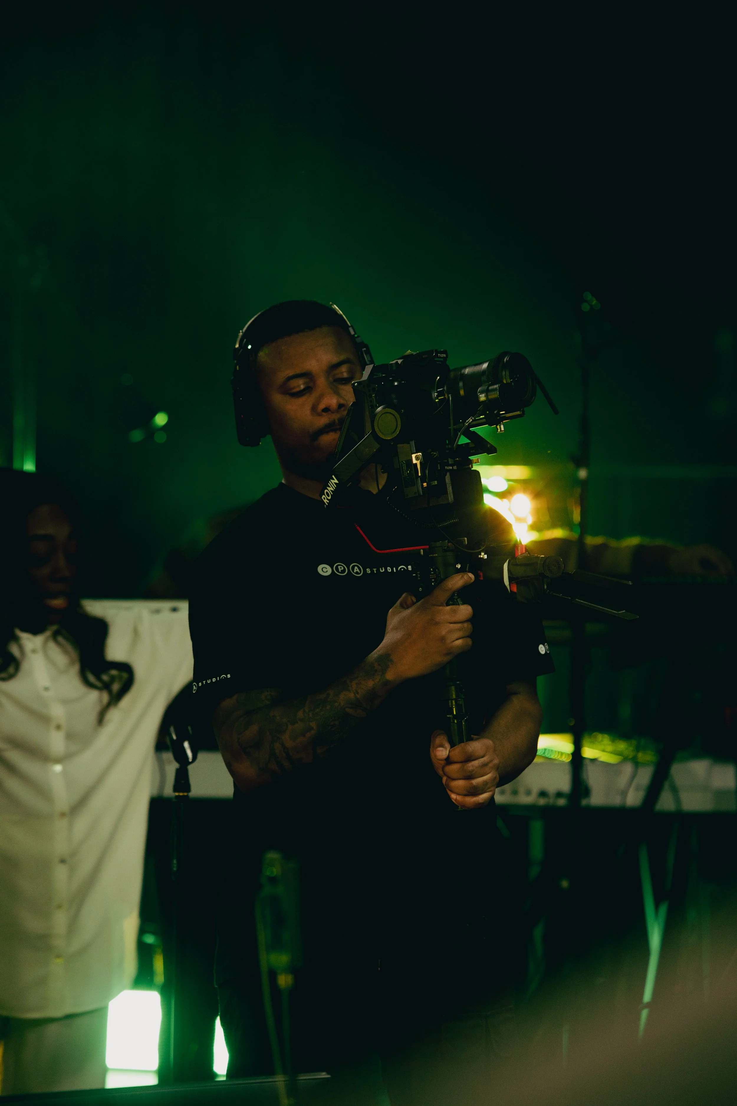 A man in a black t-shirt and headphones operating a professional camera on a tripod in a dimly lit environment with green lighting.