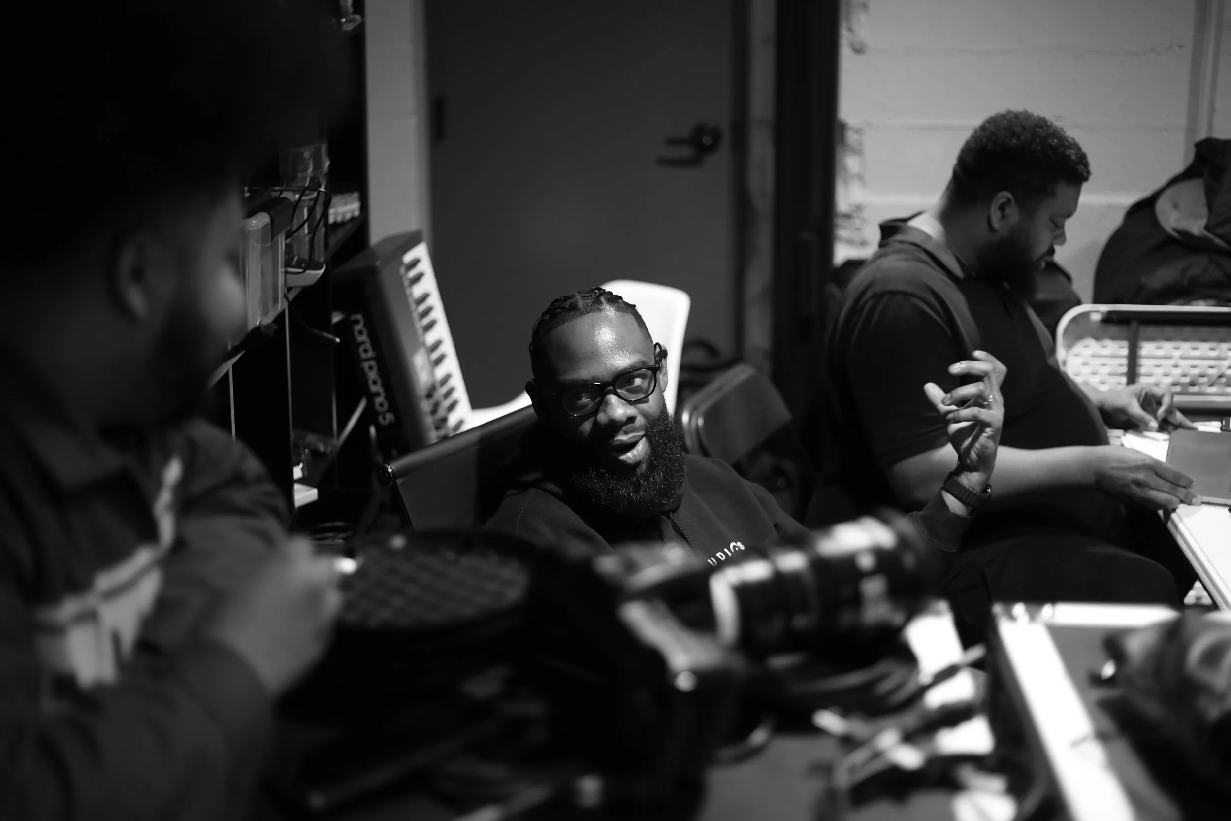 Black and white photo of four men sitting in a room, one in the foreground with glasses and braided hair, appearing to speak or sing, with DJ equipment and keyboard behind him. The other three men sit with their heads bowed or eyes closed.