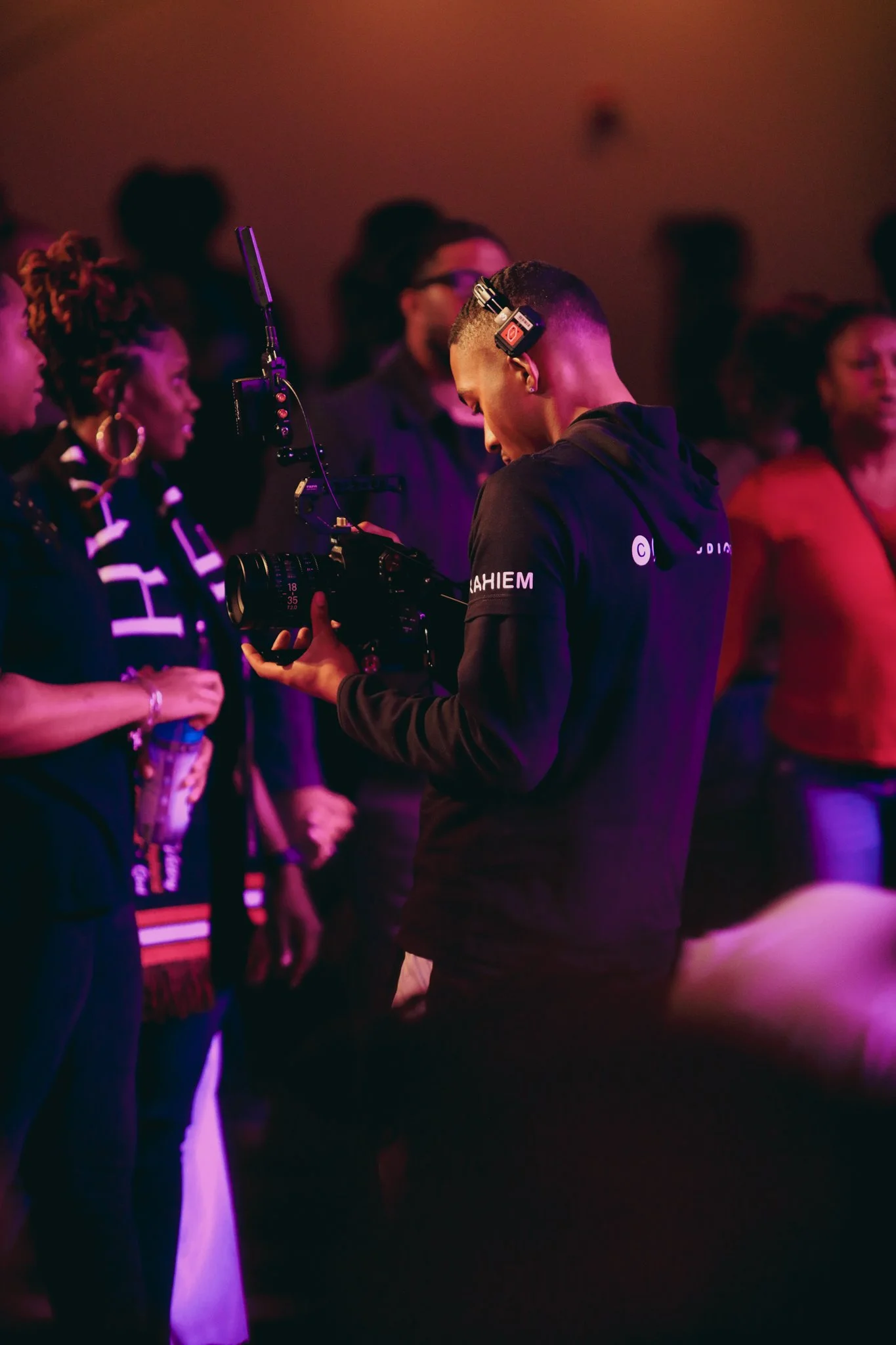 A person operating a professional video camera at an indoor event with a crowd of people, some of whom are engaged or smiling, illuminated by pink and purple lighting.