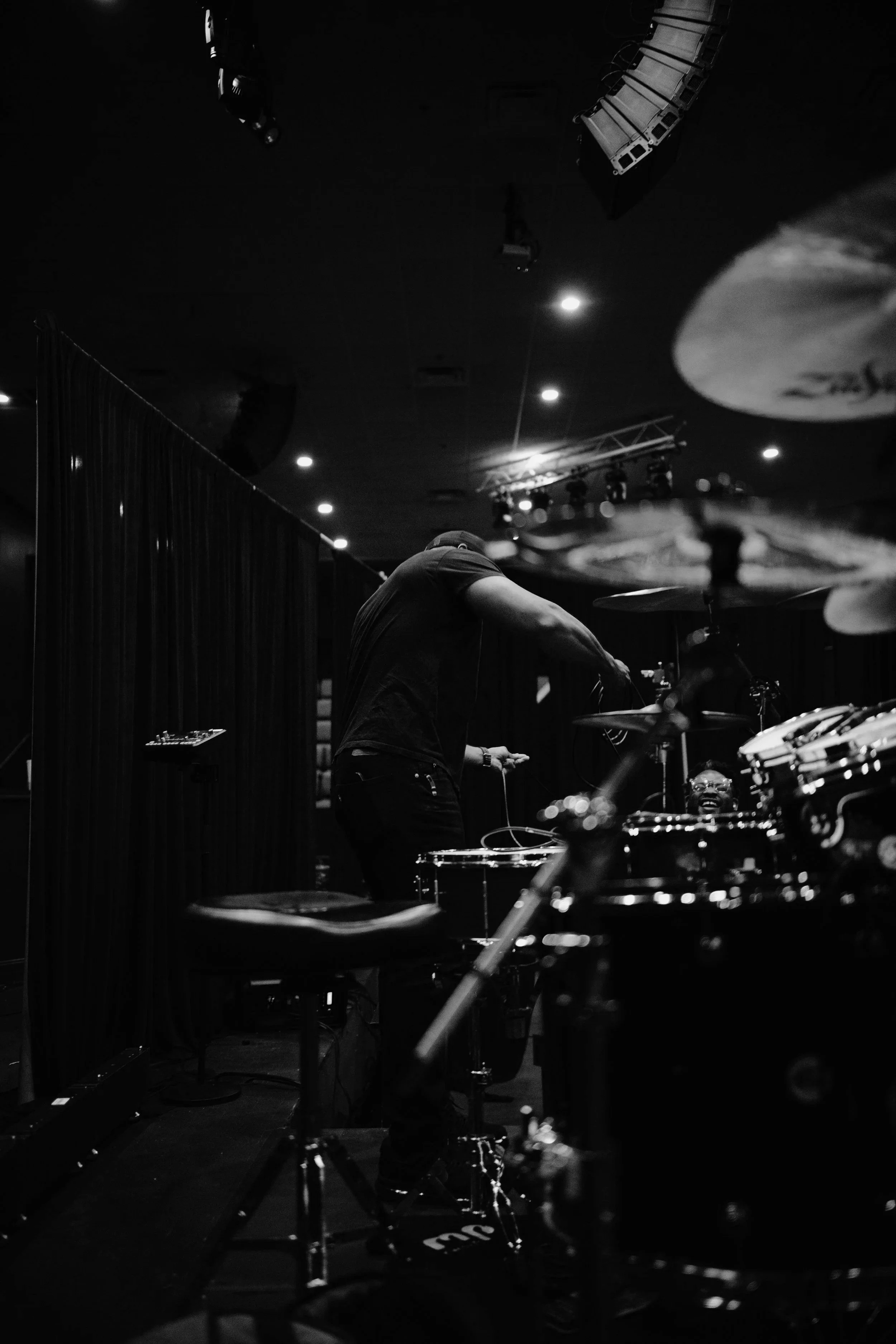 Black and white photo of a person playing drums on a stage, with instruments and stage lights visible in the background.