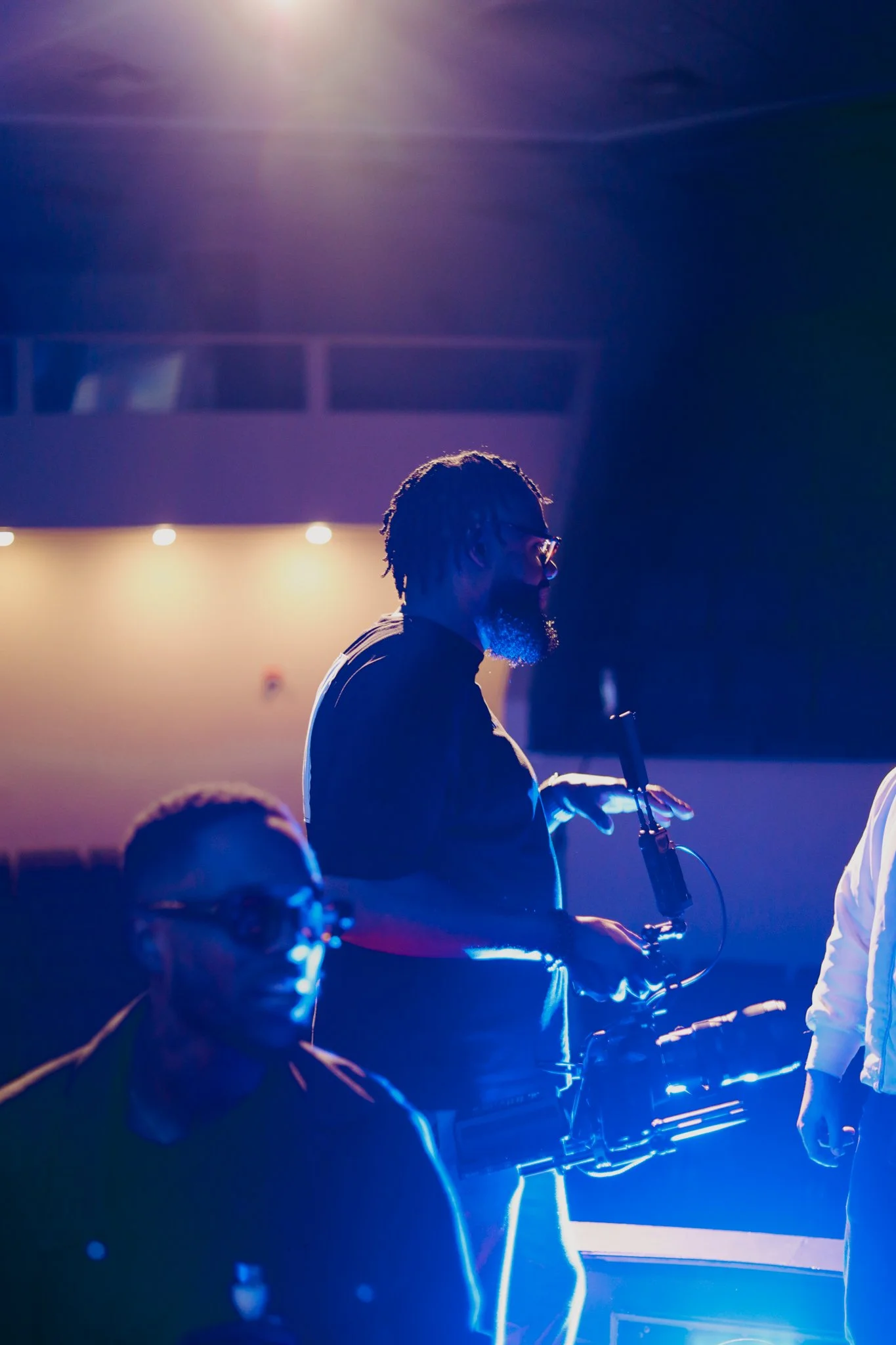 A musician playing a saxophone on stage, illuminated by colorful stage lights, with other band members partially visible in the foreground.