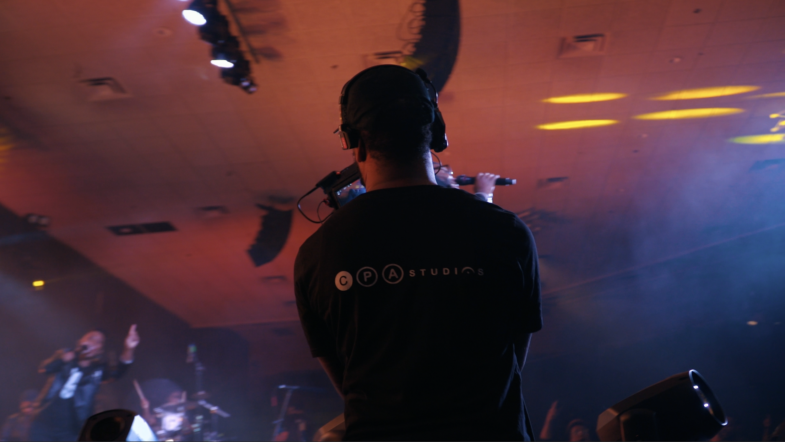 A person wearing a black shirt with the logo 'CPA STUDIOS' is standing and facing away from the camera, wearing headphones, in a dimly lit environment with colorful lights, possibly a stage or club, while holding a microphone.