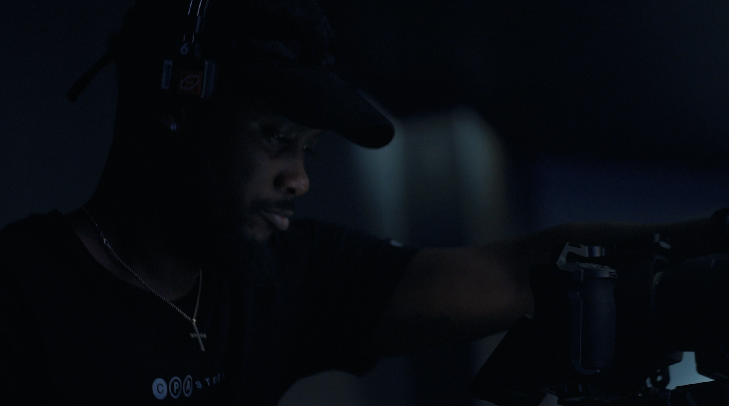 A man wearing a baseball cap and a black shirt is operating a professional microphone in a dimly lit environment, with a slight light illuminating his face.