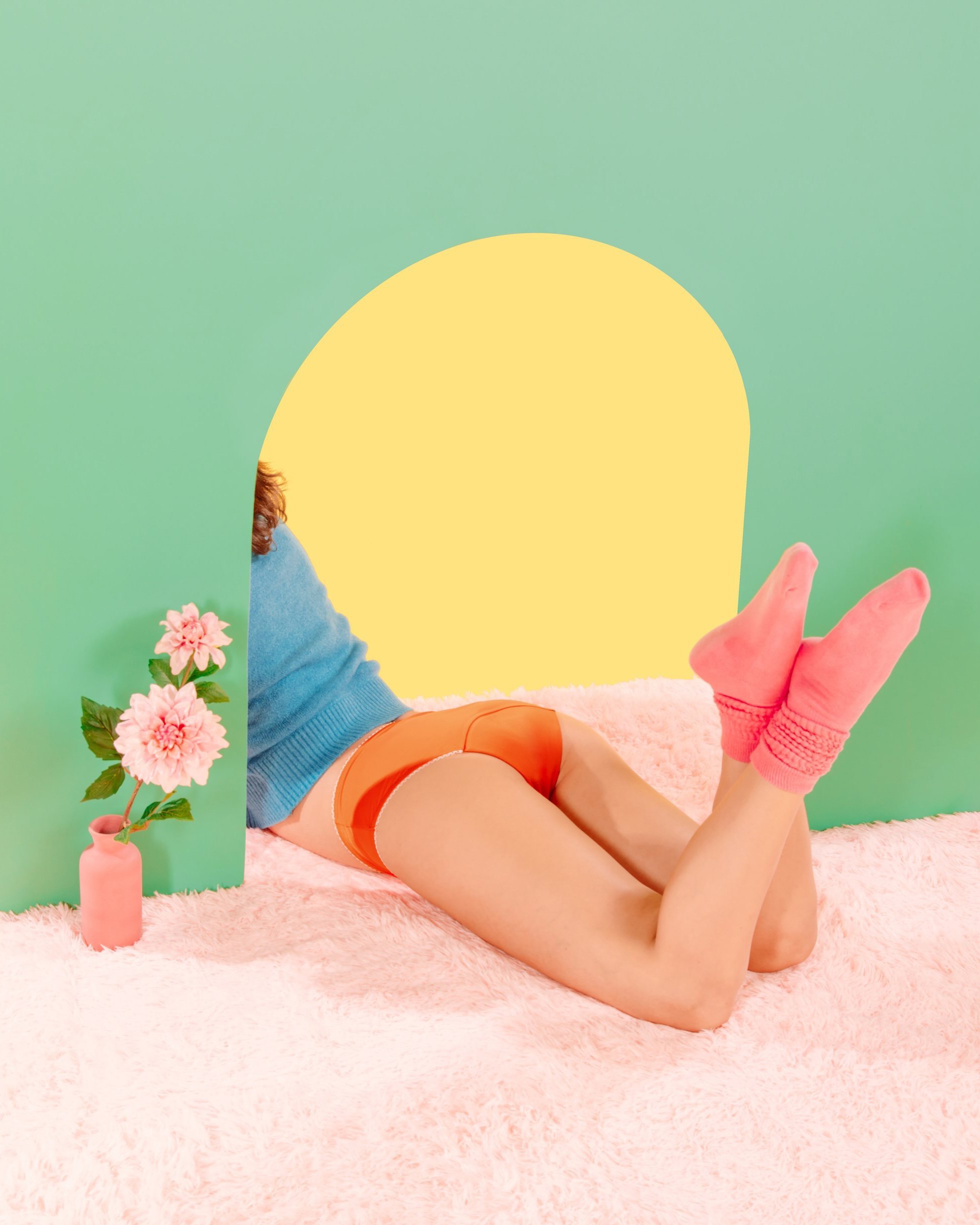 Person lying on a pink fuzzy rug, wearing orange underwear, a blue top, and pink socks, with a light green background. There is a small vase with pink flowers on the left.