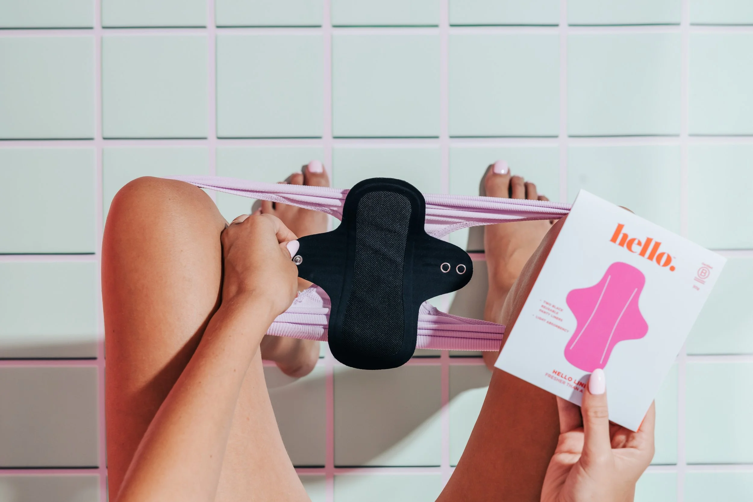 Hello Period reusable pad packaging held by woman sitting on the toilet. Pale pink nails. Blue and purple tiles. Studio photography, packaging design, branding, typography, designer. Alice Lloyd.