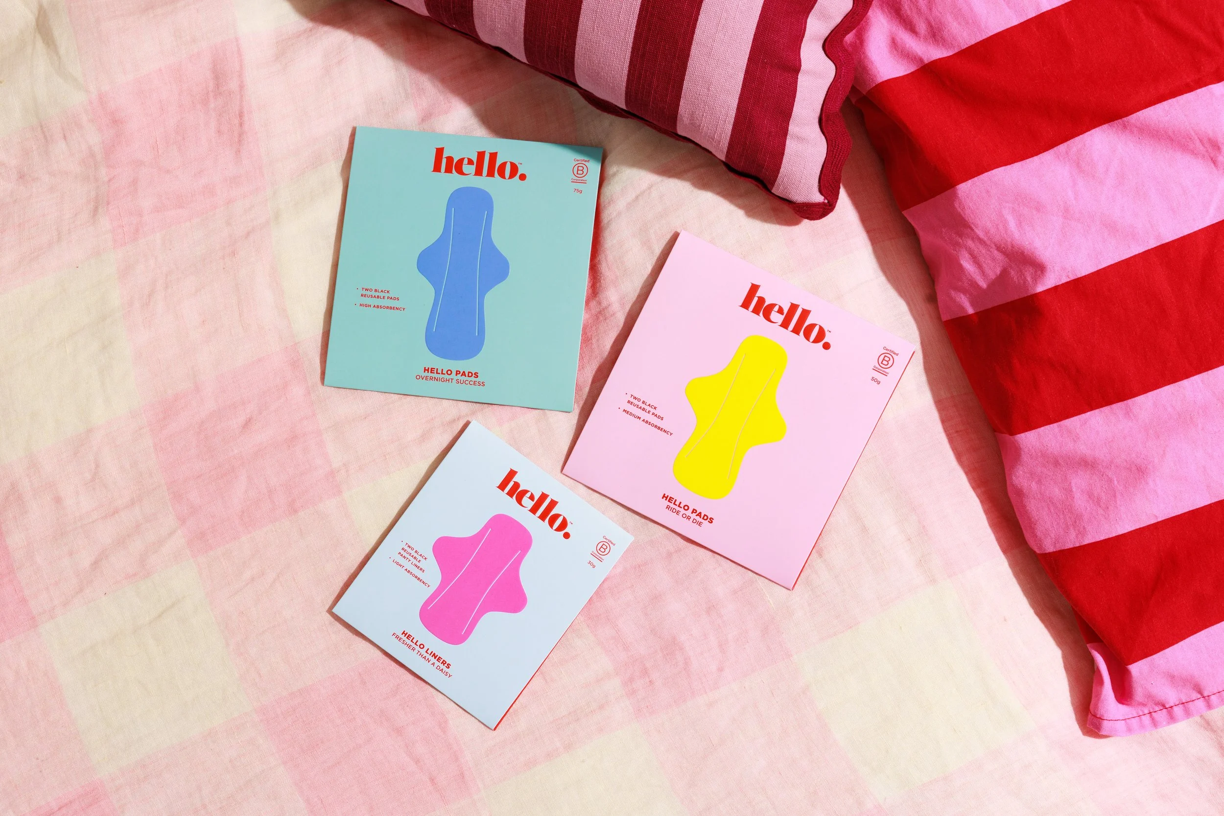 Three boxes of Hello Pads menstrual products in pink, blue, and yellow packaging, lying on a pink checkered fabric surface next to red and pink striped fabric.
