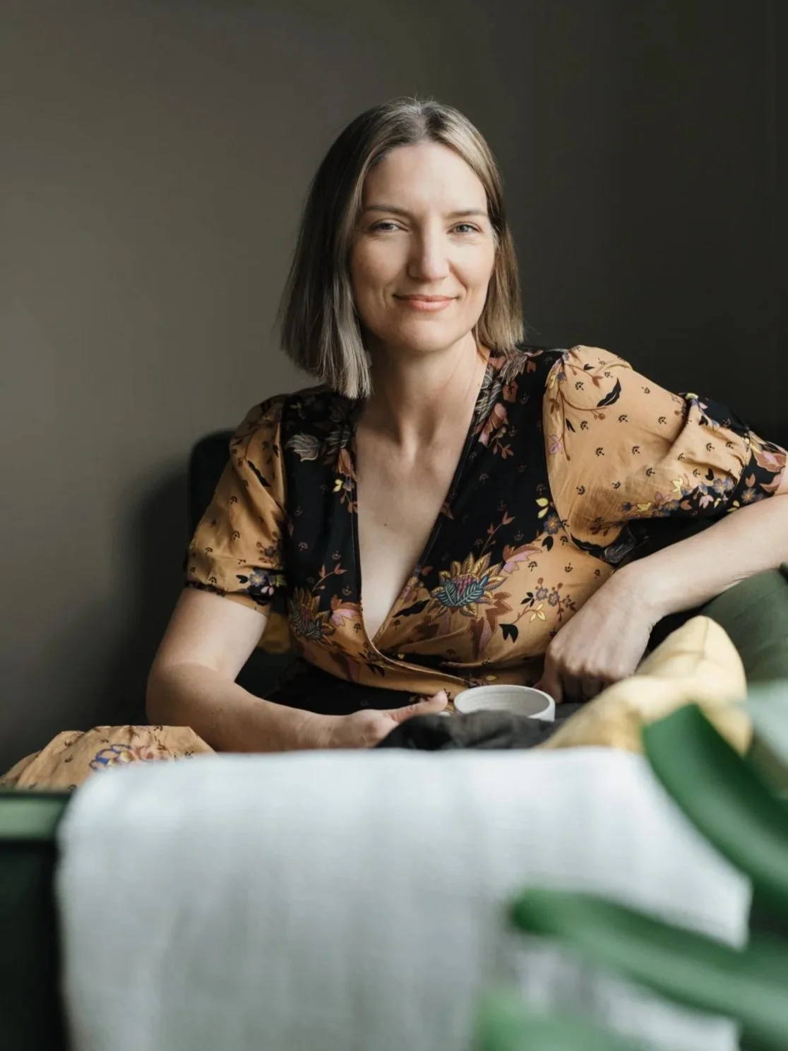 A woman with shoulder-length brown hair, wearing a floral-patterned dress, smiling and sitting on a green sofa with a cup in her hand. Designer, Creative Director, Design Studio, Wairarapa, New Zealand.