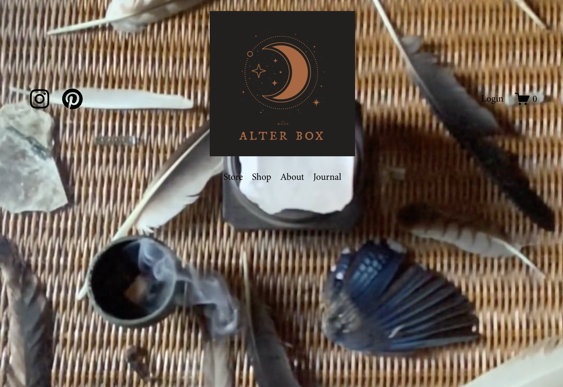 Website homepage for Alter Box e-commerce store. A table with a black and orange Moon-themed alt text image in the center, surrounded by feathers, rocks, and incense, with various icons and text overlay including social media and shopping cart icons.
