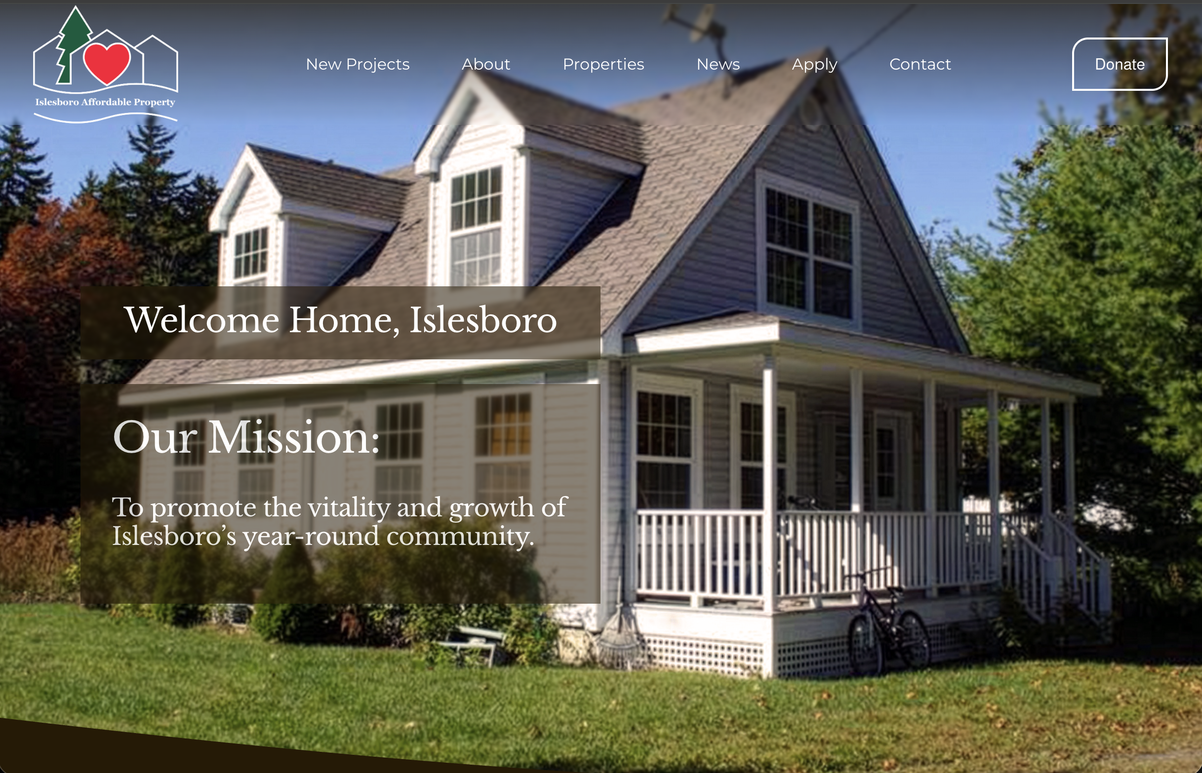 Website homepage for Islesboro Affordable Property non-profit. A two-story house with white siding and white trim, featuring large windows, a covered porch, and a front yard with green grass, trees, and shrubbery. The sky is clear and blue.