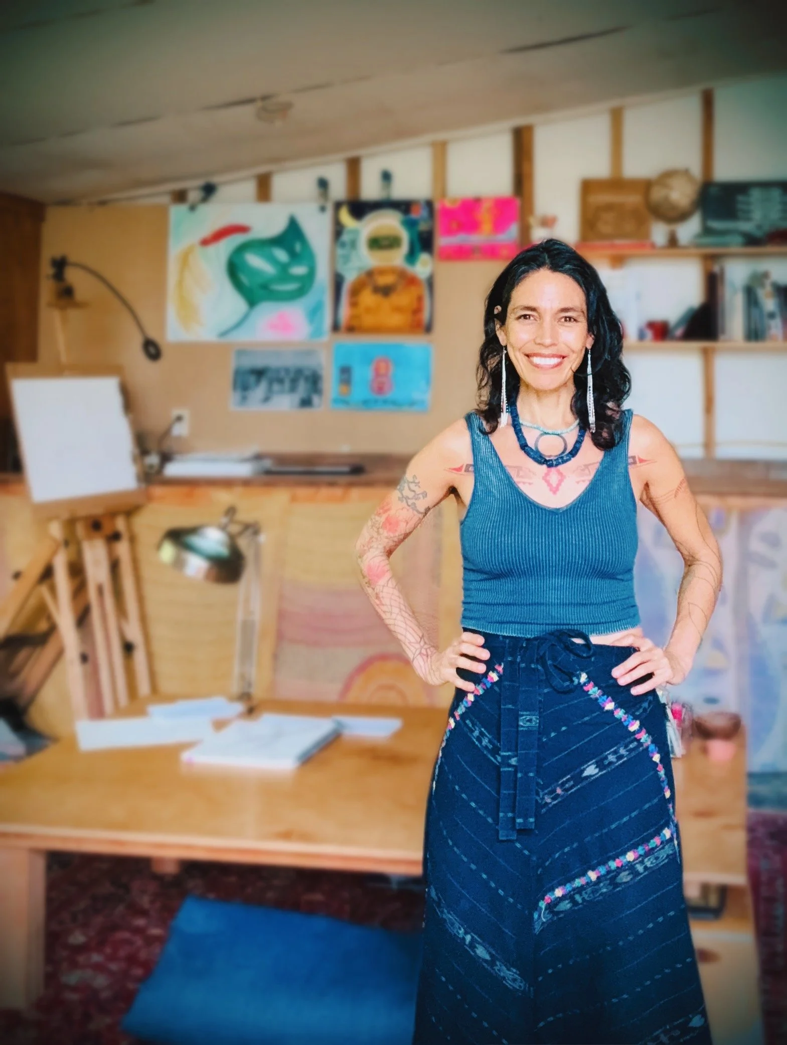 A smiling woman with dark hair and tattoos wearing a blue top and a patterned skirt standing in an art studio with paintings on the wall behind her.
