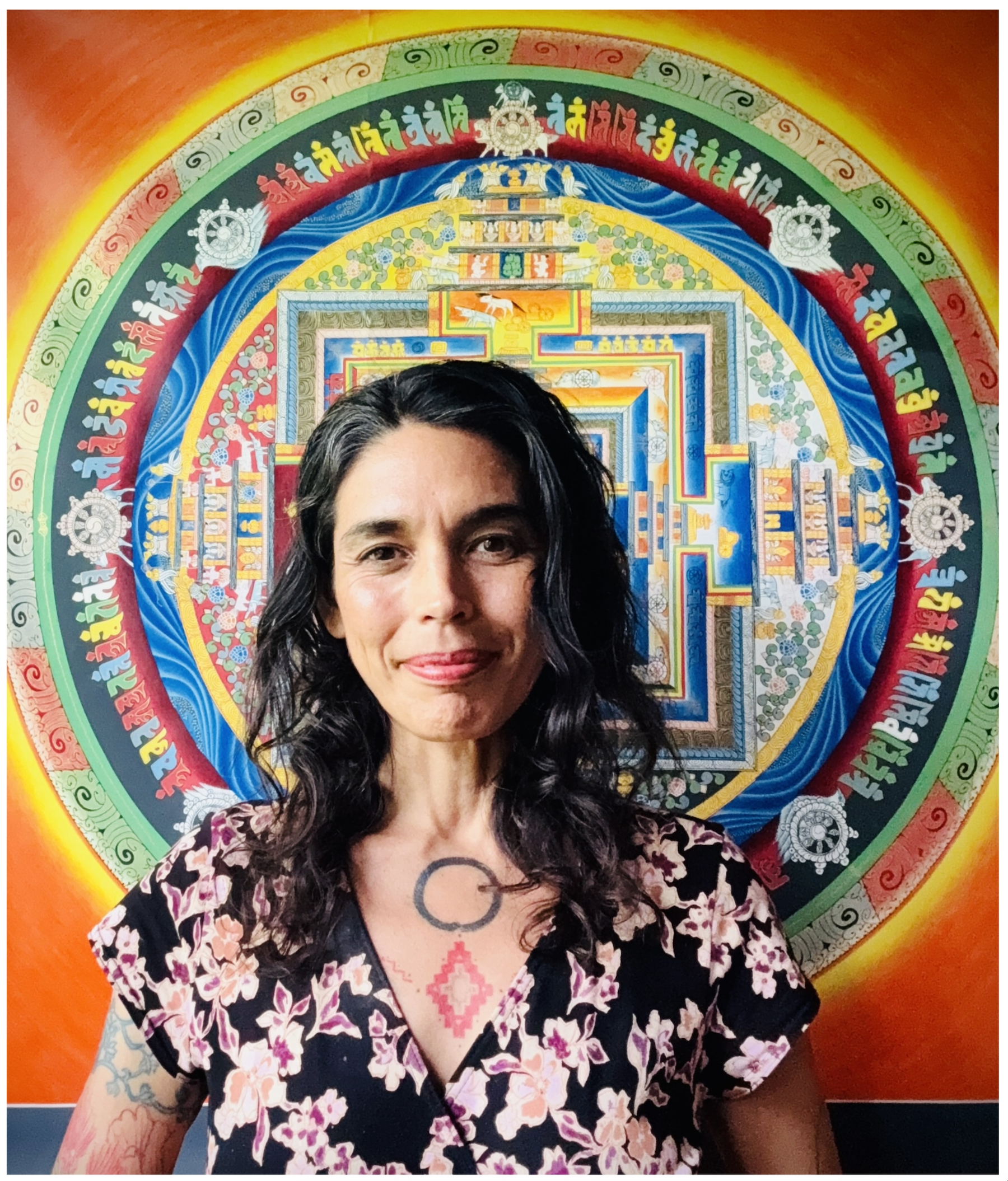 A woman with dark hair and tattoos on her arm standing in front of a colorful, intricate mural featuring a spiral pattern with vibrant colors and detailed symbols and designs.