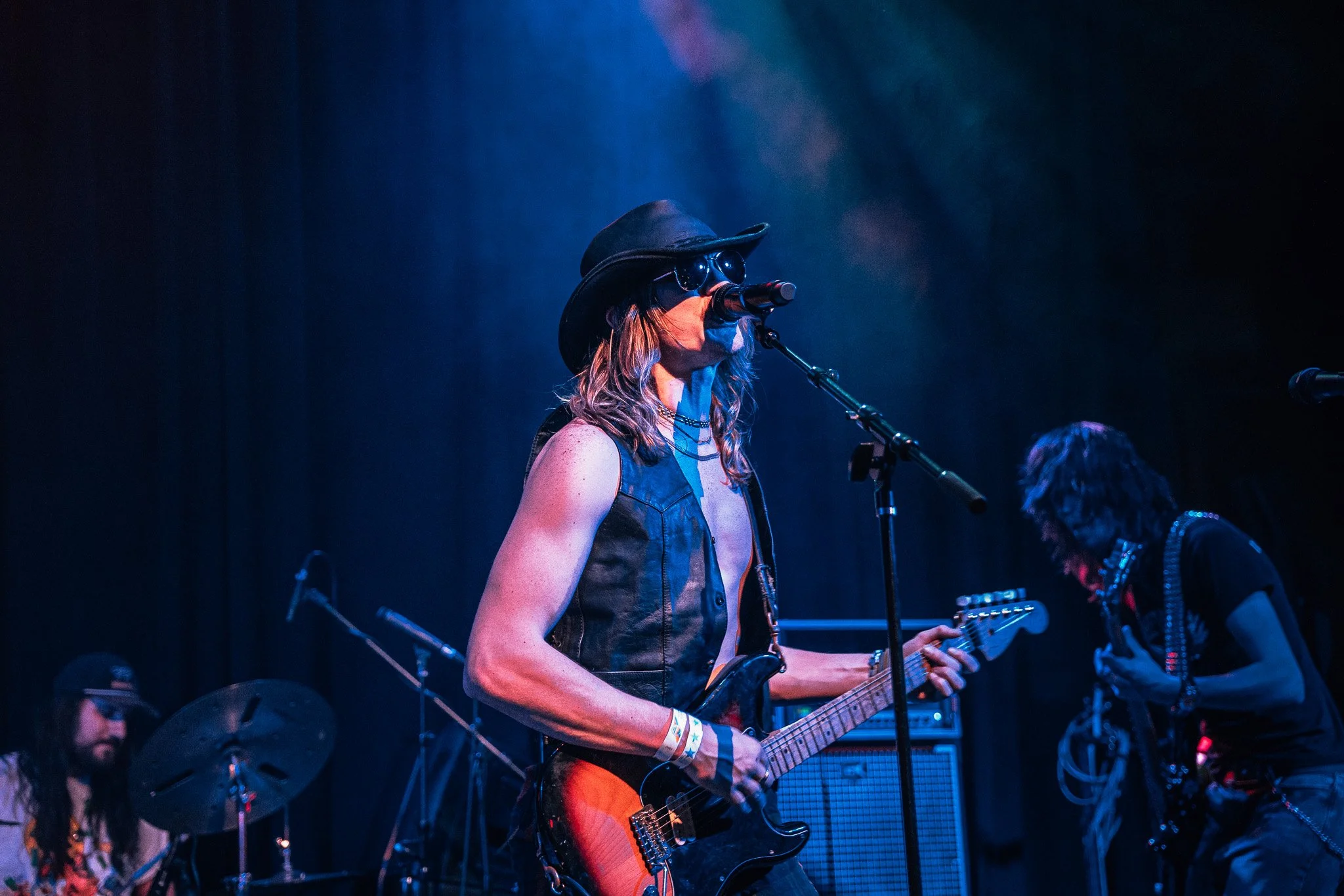 A female musician performs on stage playing an electric guitar and singing into a microphone. She wears a wide-brimmed hat, sunglasses, and a sleeveless top. There are other band members visible in the background with musical equipment and stage lighting creating a blue tone.
