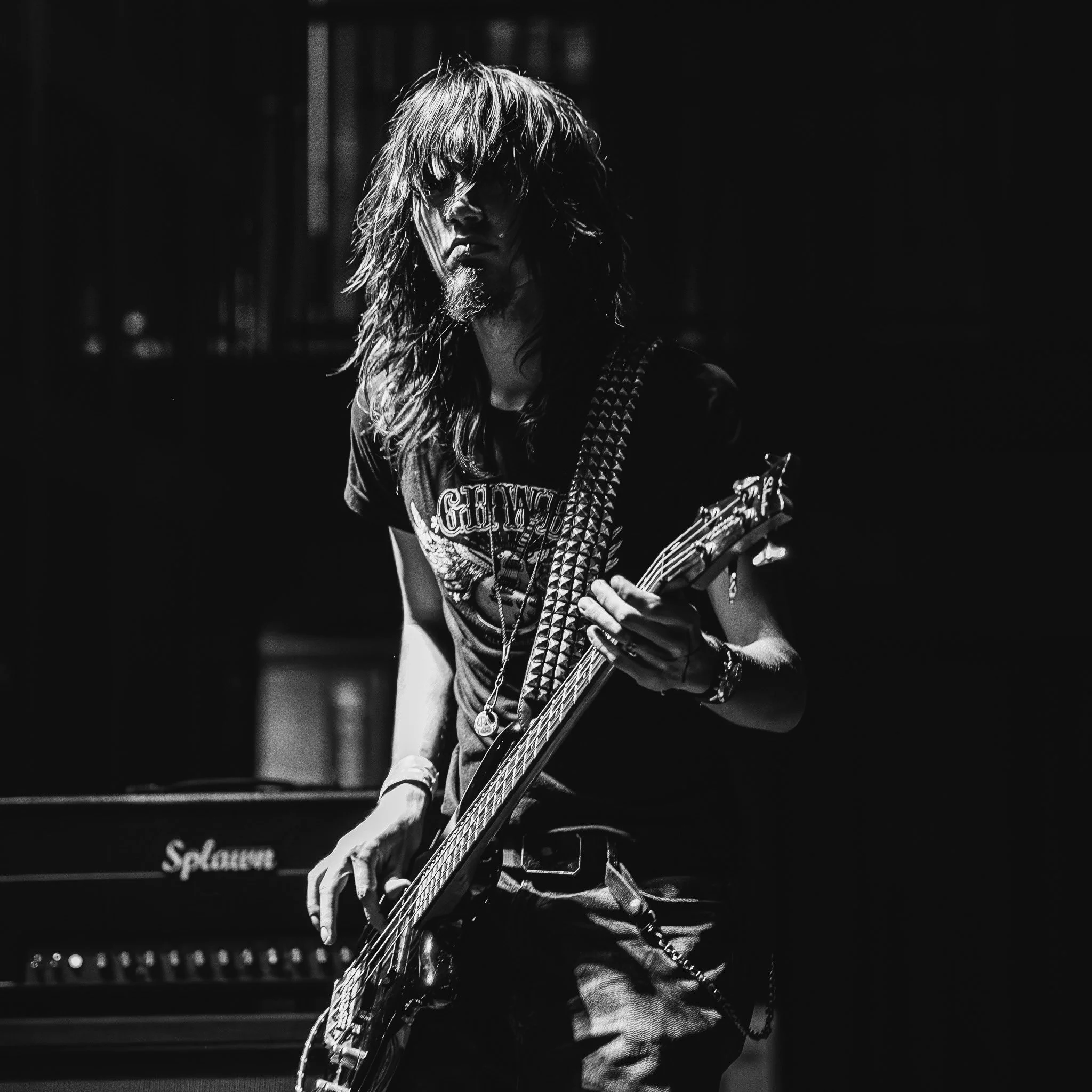 A black and white photo of a long-haired male guitarist, with a beard and wearing a T-shirt, playing an electric guitar during a live performance.