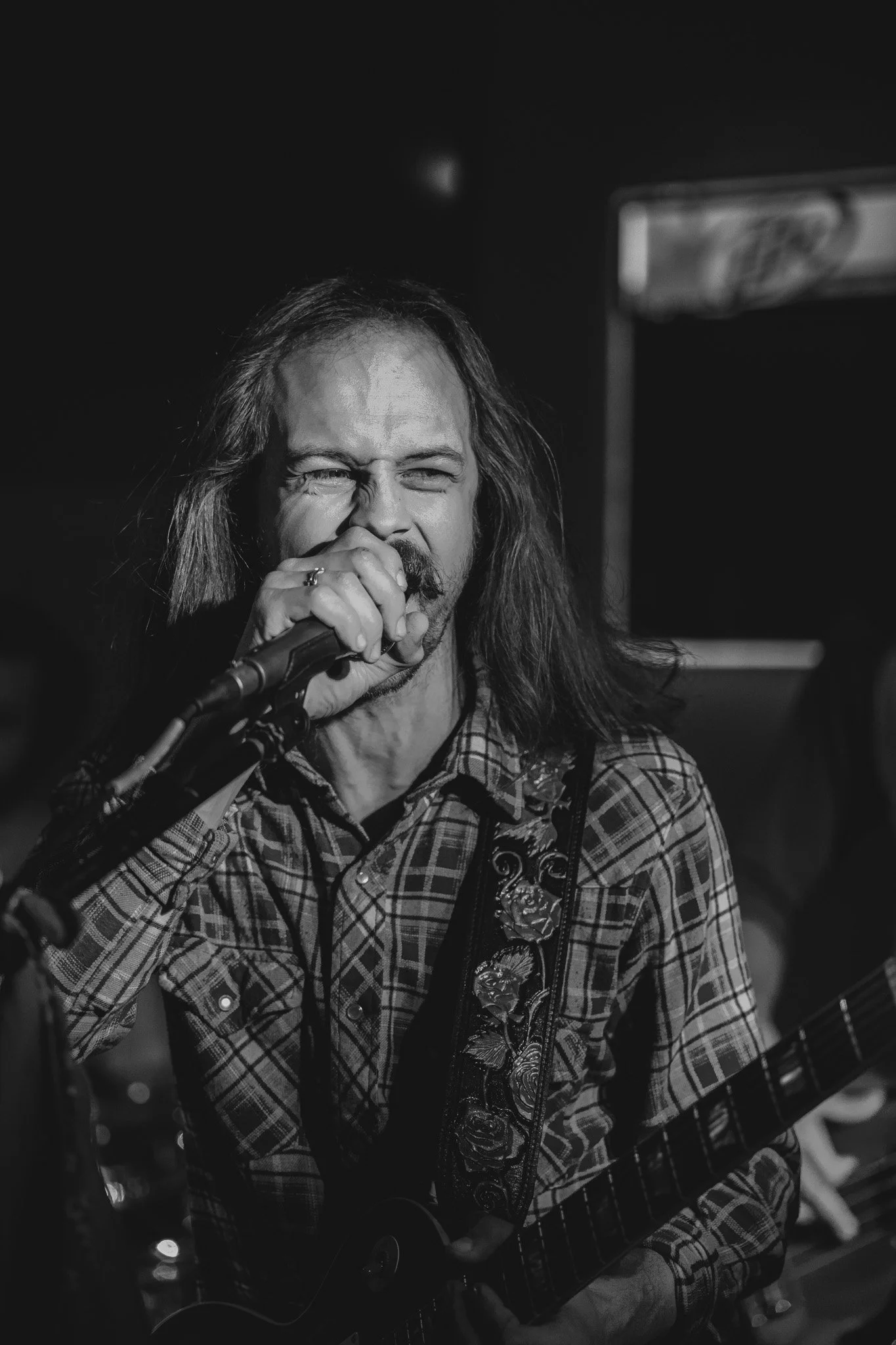 A man with long hair and a mustache singing into a microphone while playing an acoustic guitar in a dimly lit setting.